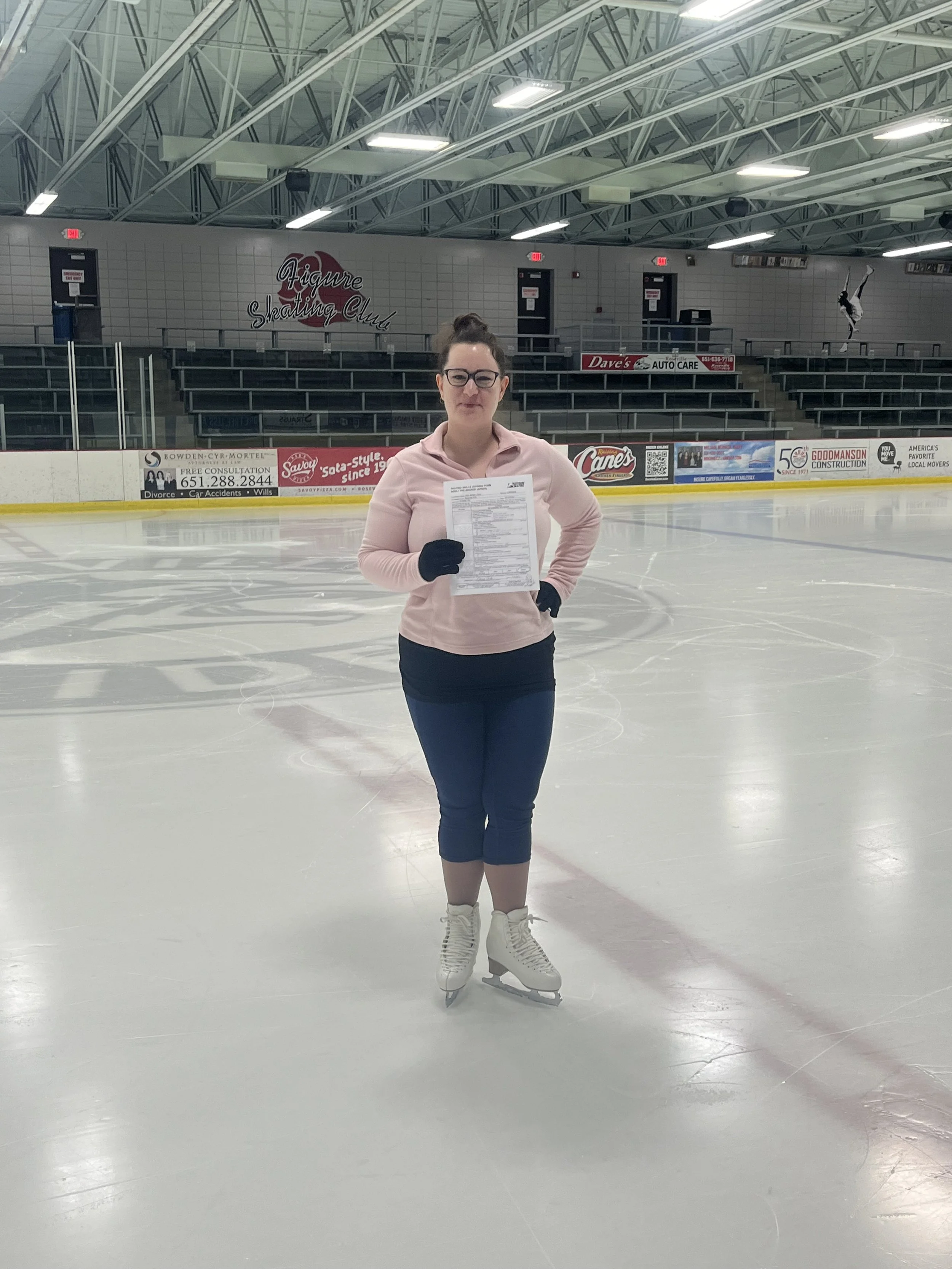 A woman in ice skates holding a piece of paper on an ice rink, with empty bleachers and banners in the background.