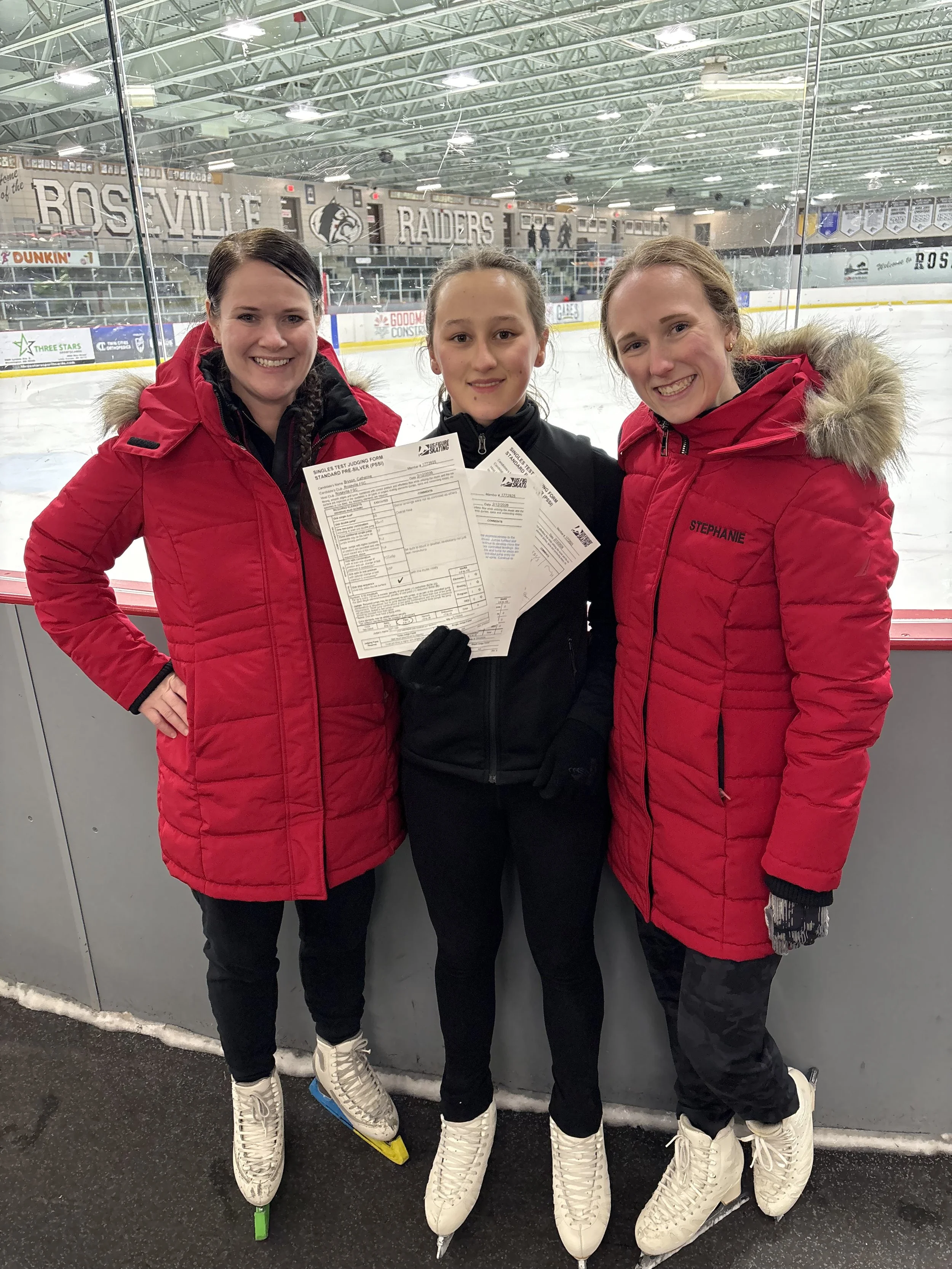 Three young women standing on ice skates at an indoor ice rink, smiling and holding papers, with the rink and banners in the background.