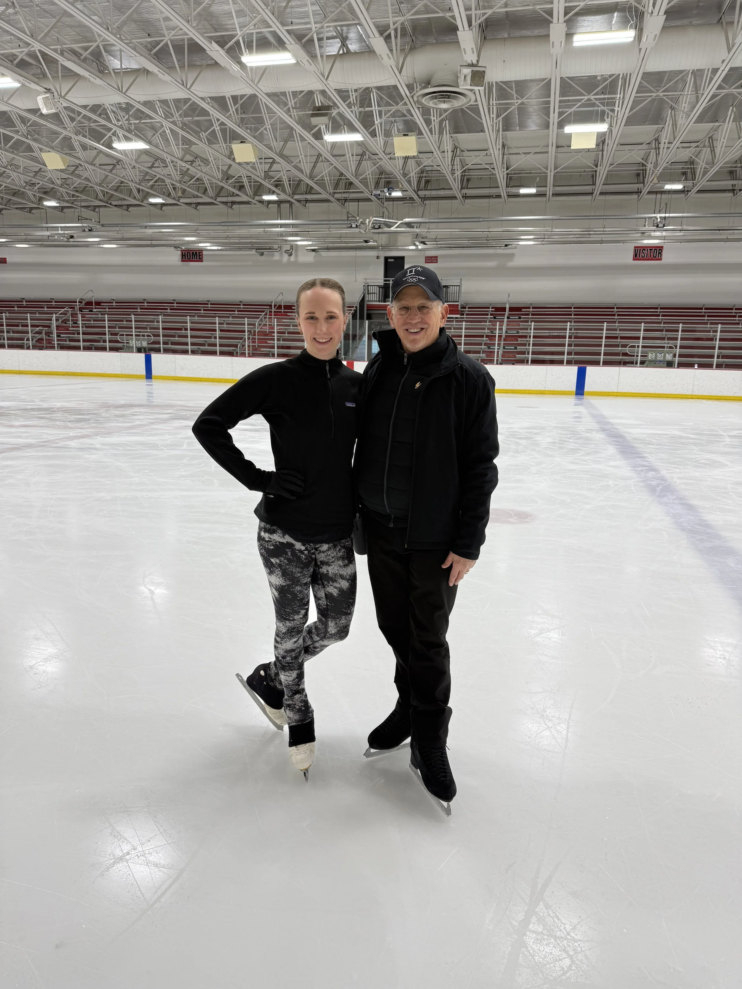 Two people standing on an ice skating rink, smiling at the camera, with an empty stadium stands background.