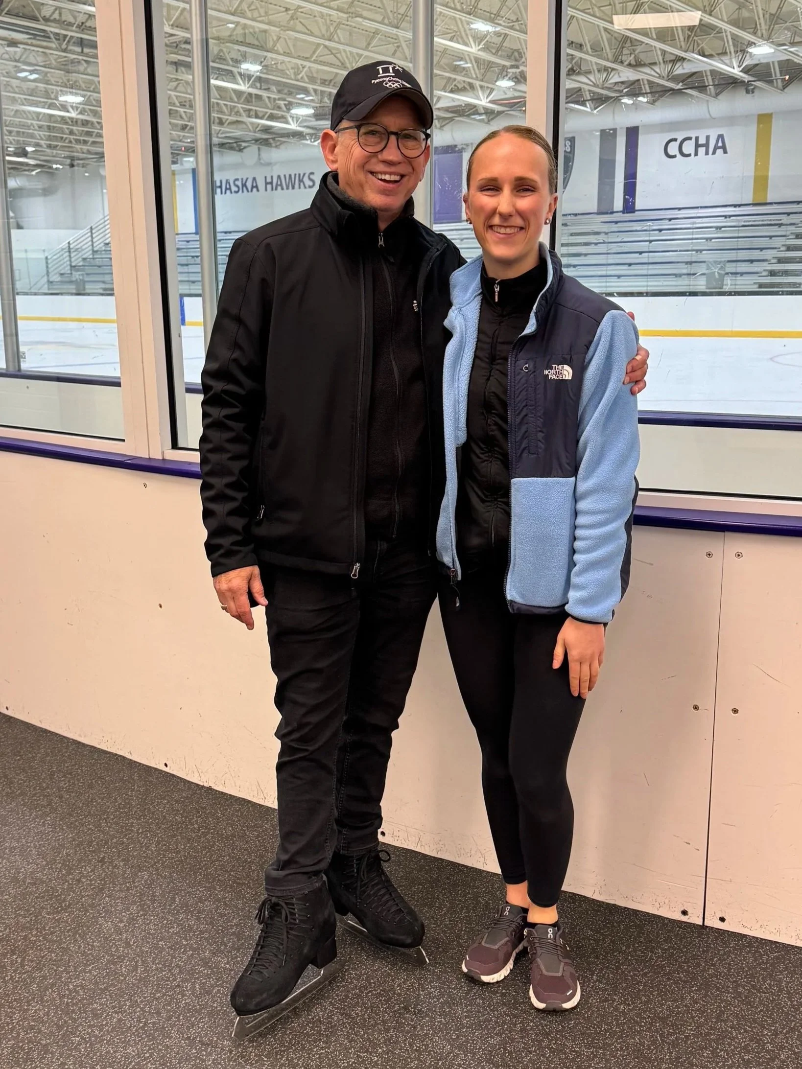 A man and a woman in athletic wear standing inside an ice skating rink, smiling and posing for a photo.
