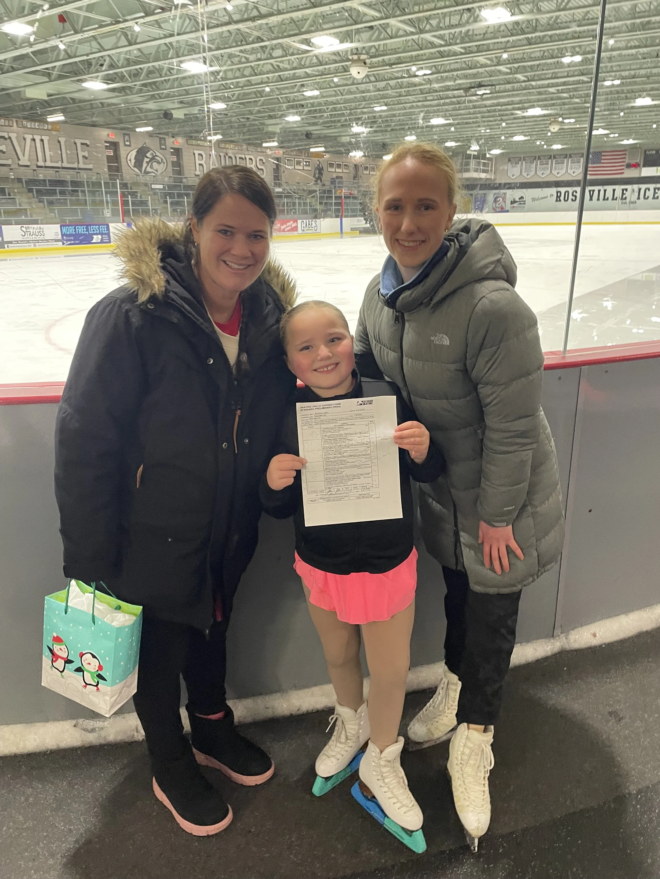 A young girl in skating gear holding a form, flanked by two women, at an indoor ice rink.