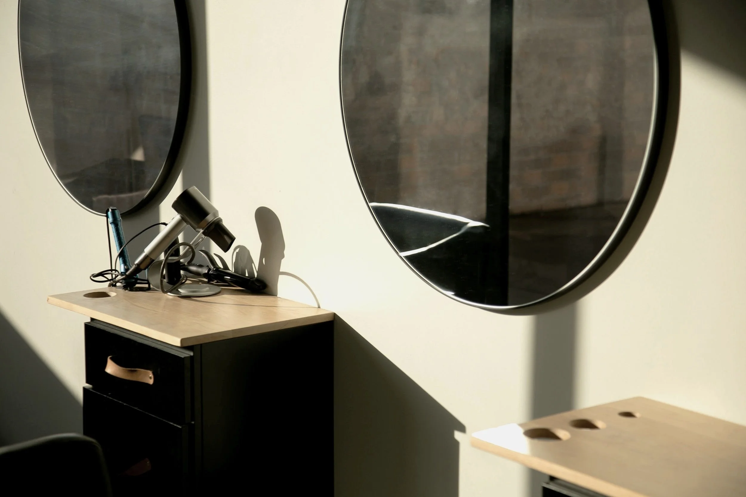 Interior scene with two large round mirrors on a beige wall, a black cabinet with a light wood top, and a cluster of hairstyling tools including a hairdryer and a round comb on the wood surface. Sunlight casts shadows on the wall and furniture.