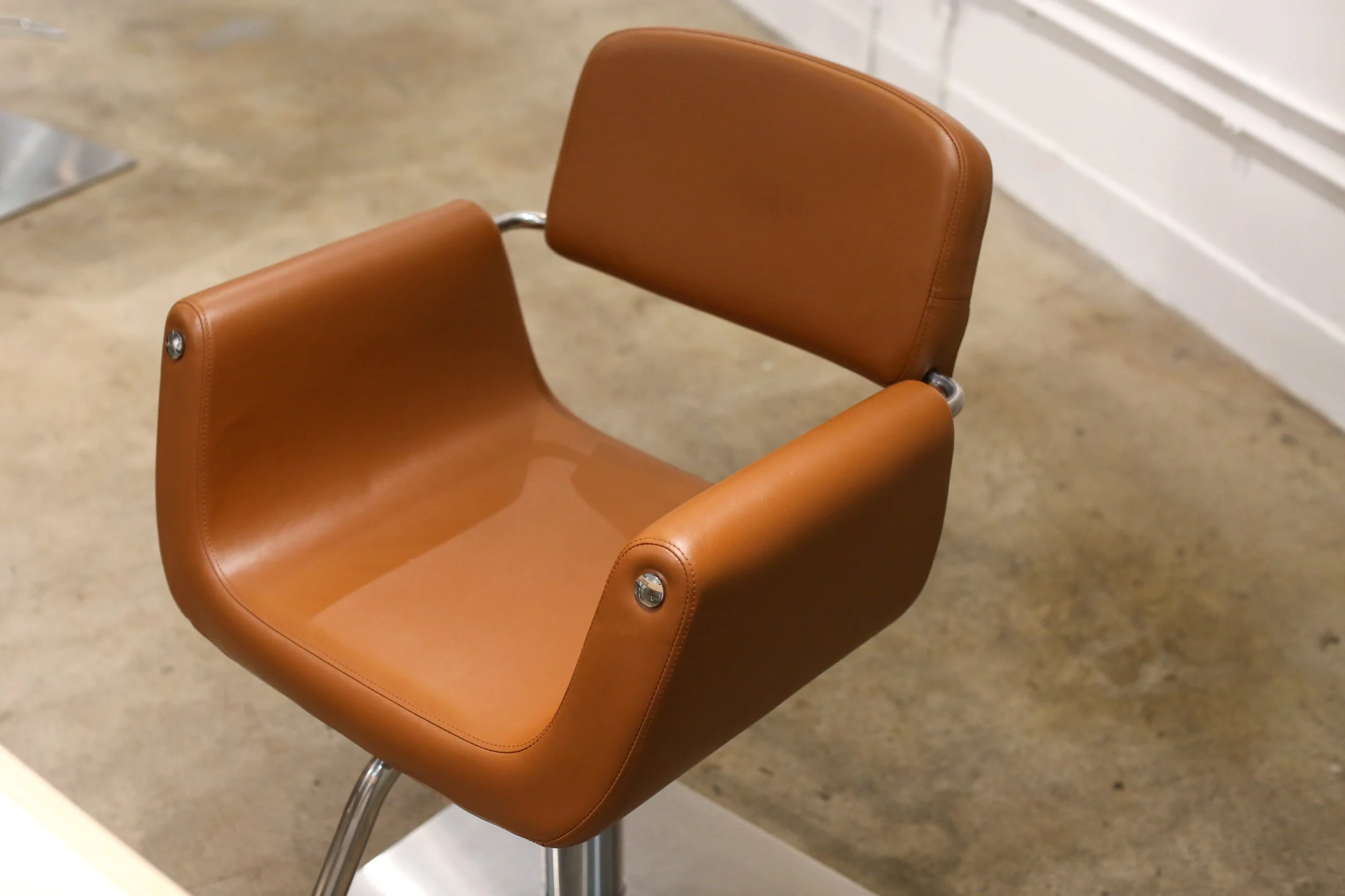 A brown leather chair with metal armrests and legs placed on a concrete floor near a white wall.