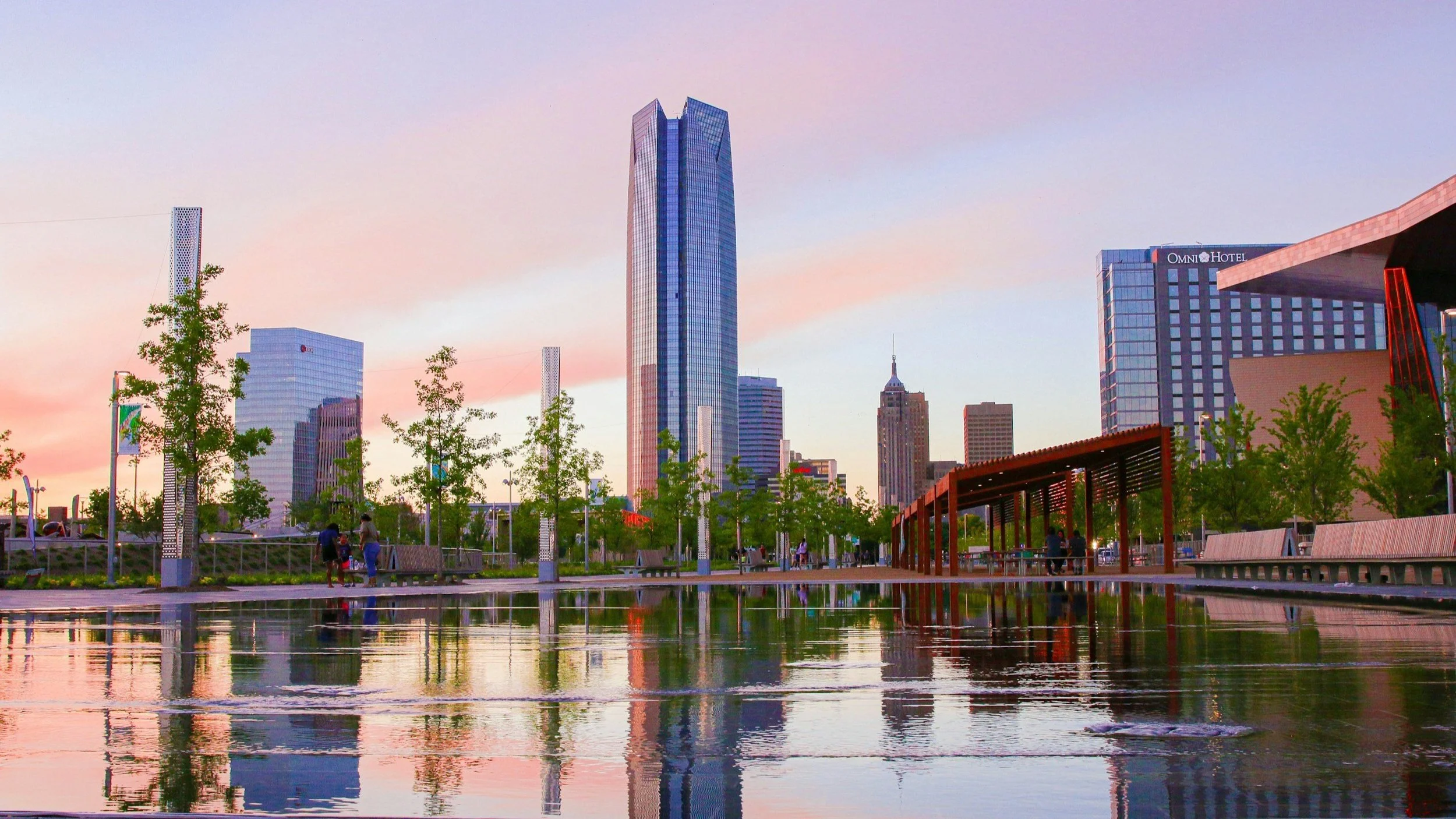 Skyline of Oklahoma City at dusk with buildings reflected in a water feature at Scissortail Park.