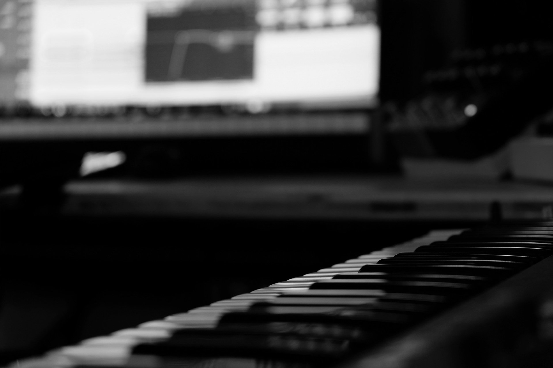 Schwarz-weiß-Bild eines Klaviers mit Blick auf die Tastatur im Vordergrund und einen Bildschirm im Hintergrund.