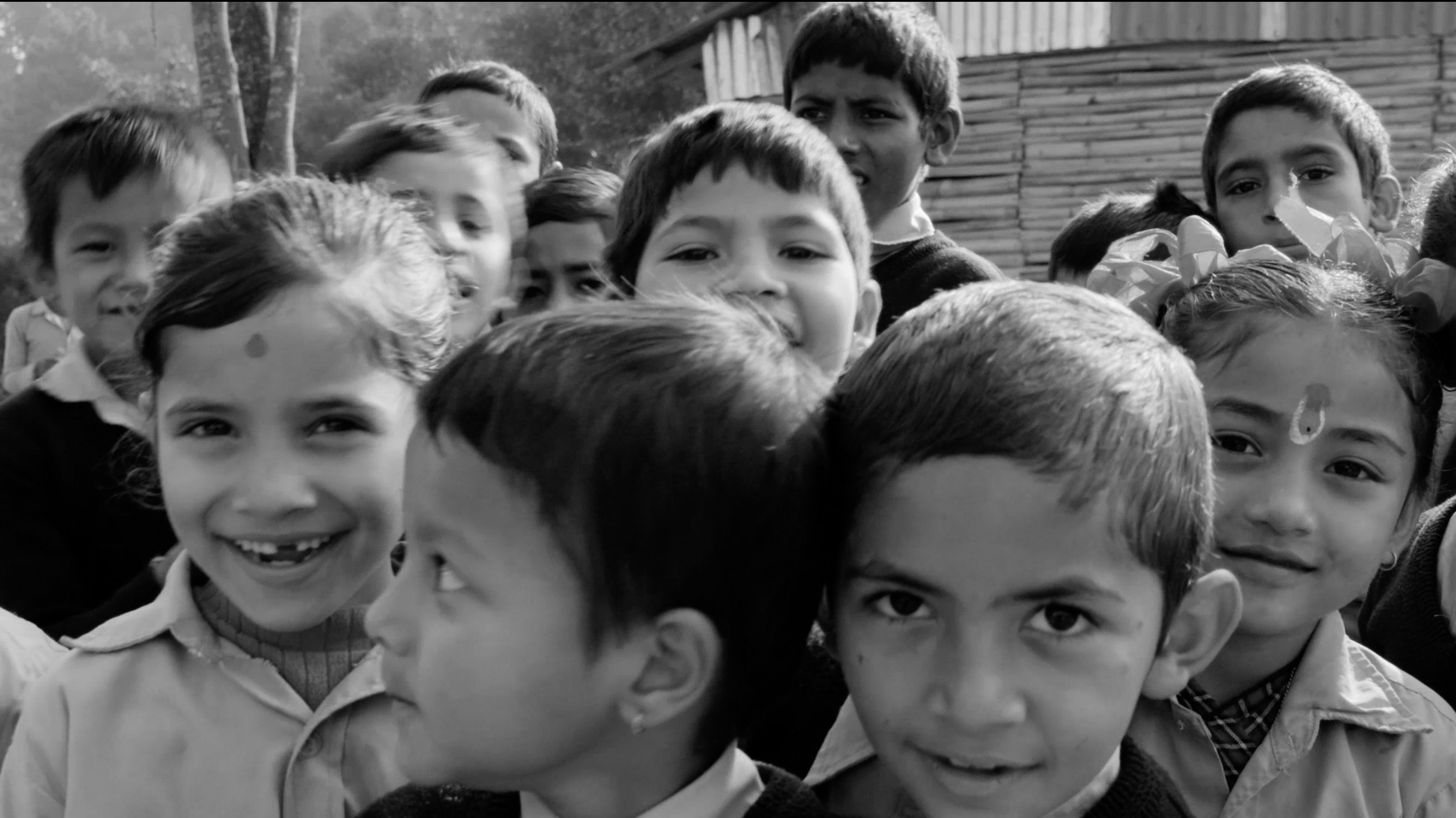 Eine Gruppe von Kindern verschiedener ethnischer Hintergründe, die draußen zusammenstehen, einige lächeln, einige schauen neugierig, eine Frau mit Schleifen im Haar im Hintergrund.