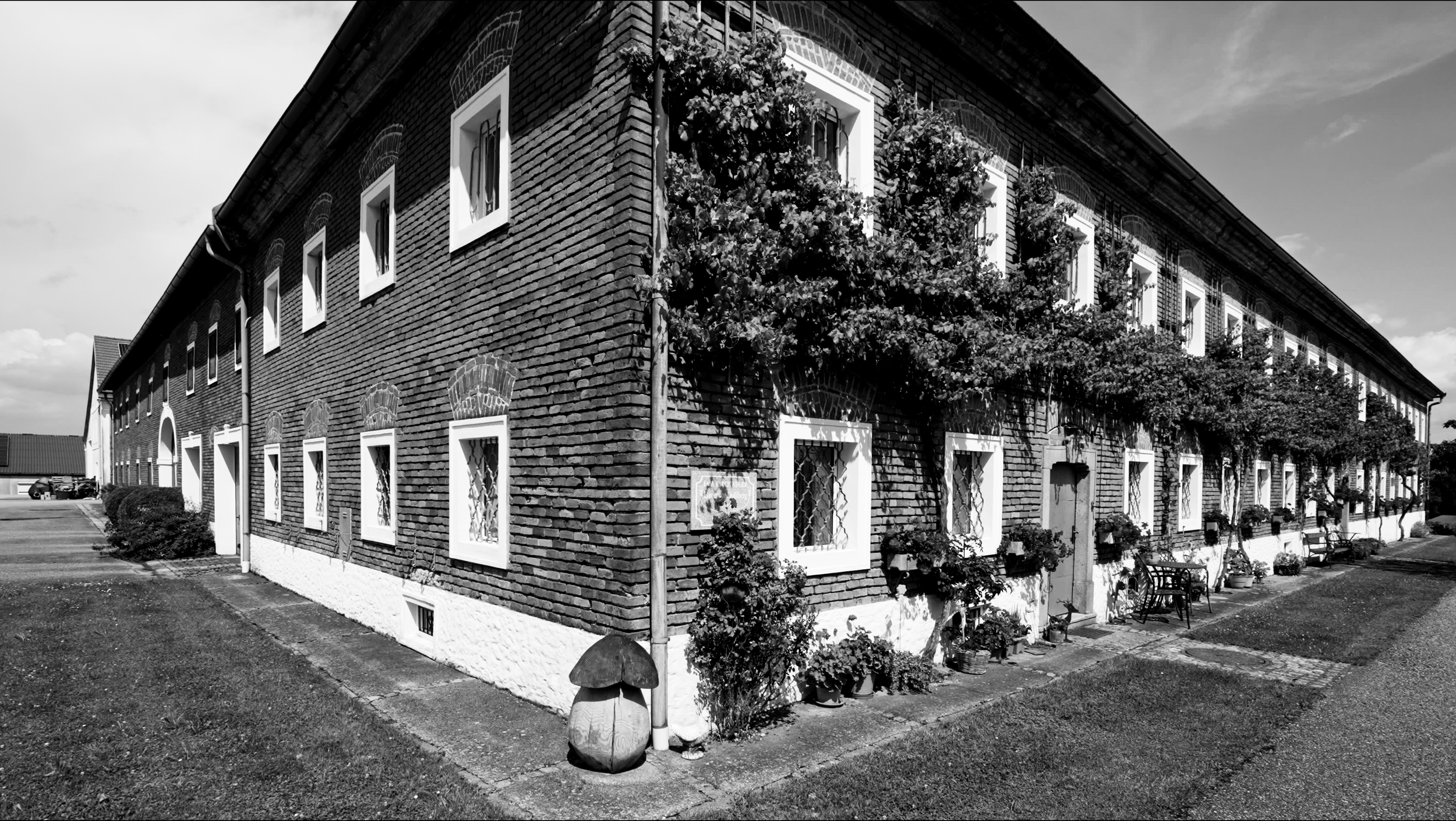 Schwarz-weiß-Fotografie eines langen, zweistöckigen Backsteinhauses mit weißen Fensterrahmen, das von Blumen und Gartenmöbeln im Vorgarten geschmückt ist.