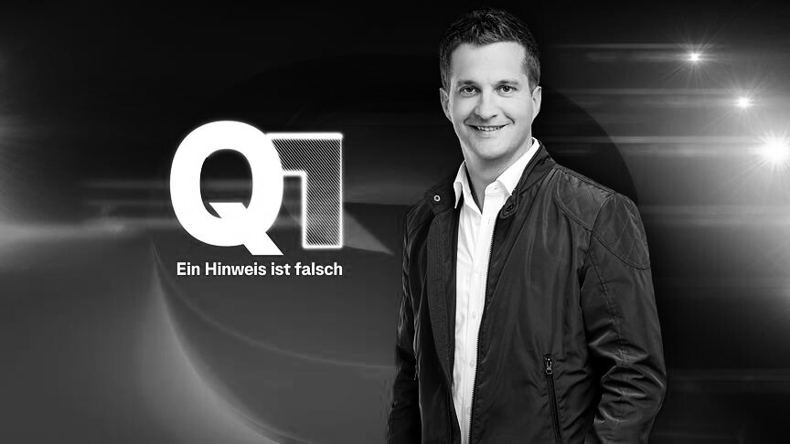 Ein Mann steht vor einem Hintergrund mit dem großen Q1-Logo und dem Text 'Ein Hinweis ist falsch'.