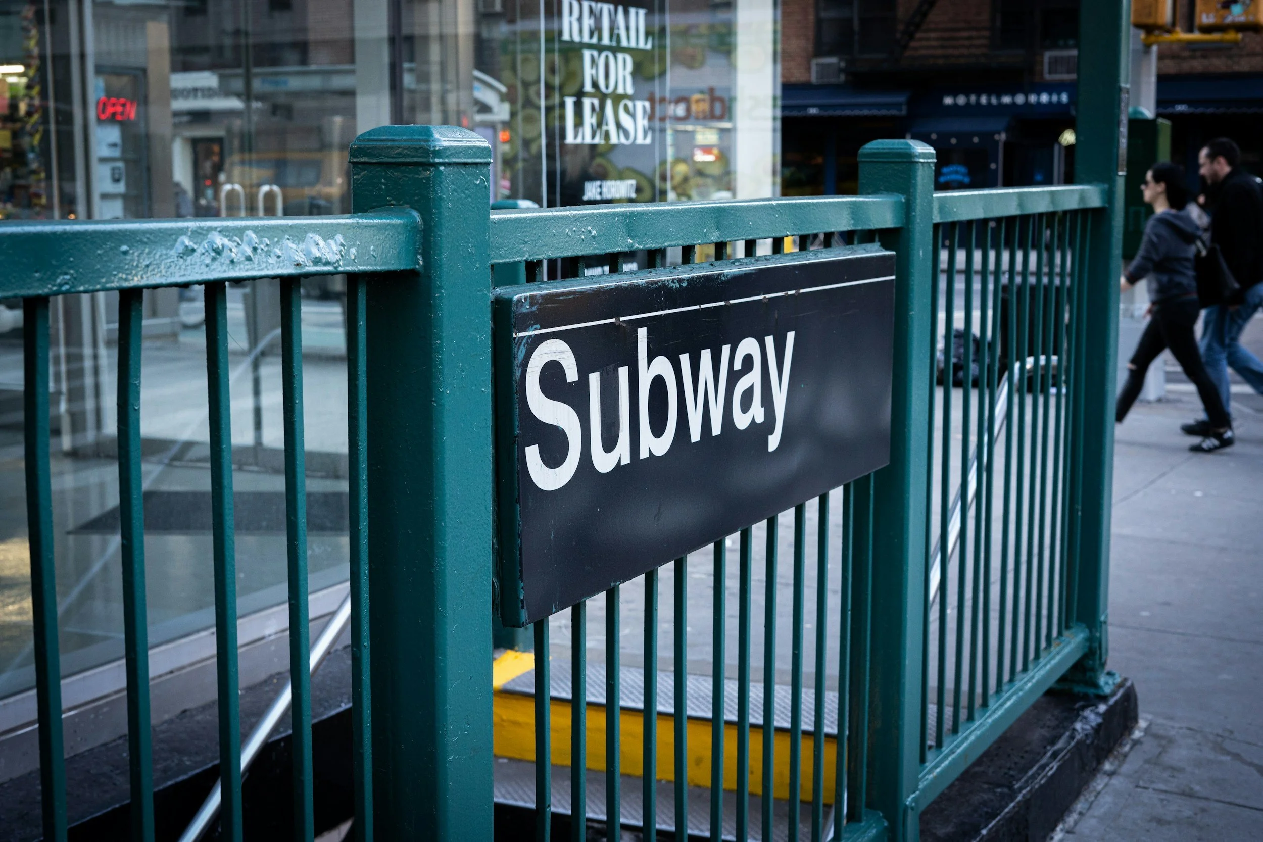 Entrance to a subway station with a sign that says 'Subway' and a background showing 'Retail For Lease' signs.