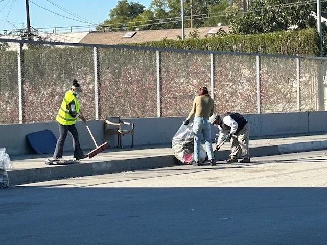 Three people cleaning a city sidewalk; one person is sweeping with a broom, and two others are picking up trash and placing it in a plastic bag.