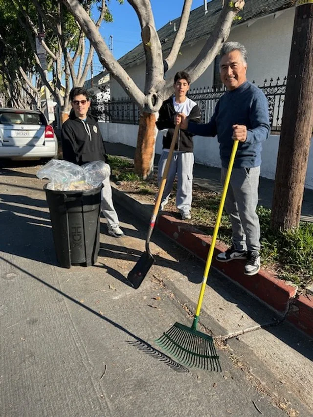 Three people cleaning a sidewalk, one man holding a rake, a teen with a garbage picker, and a woman standing next to a trash bin filled with trash bags, outdoors on a sunny day.