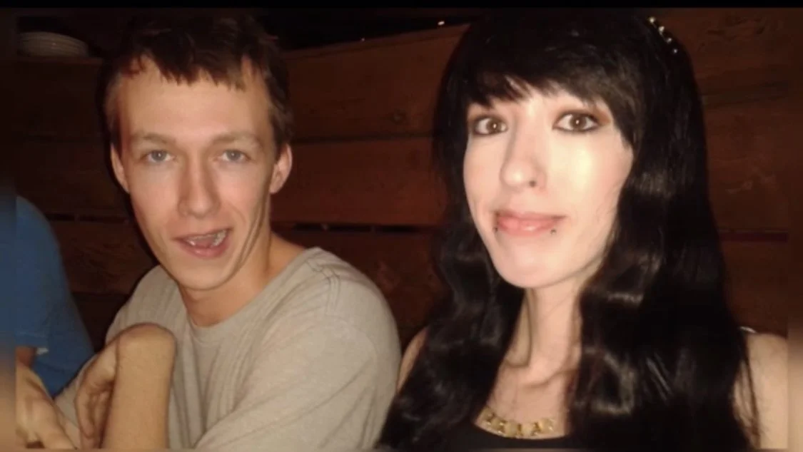 Two people posing together indoors, smiling at the camera. One is wearing a light-colored shirt, the other has long, dark hair and a necklace.