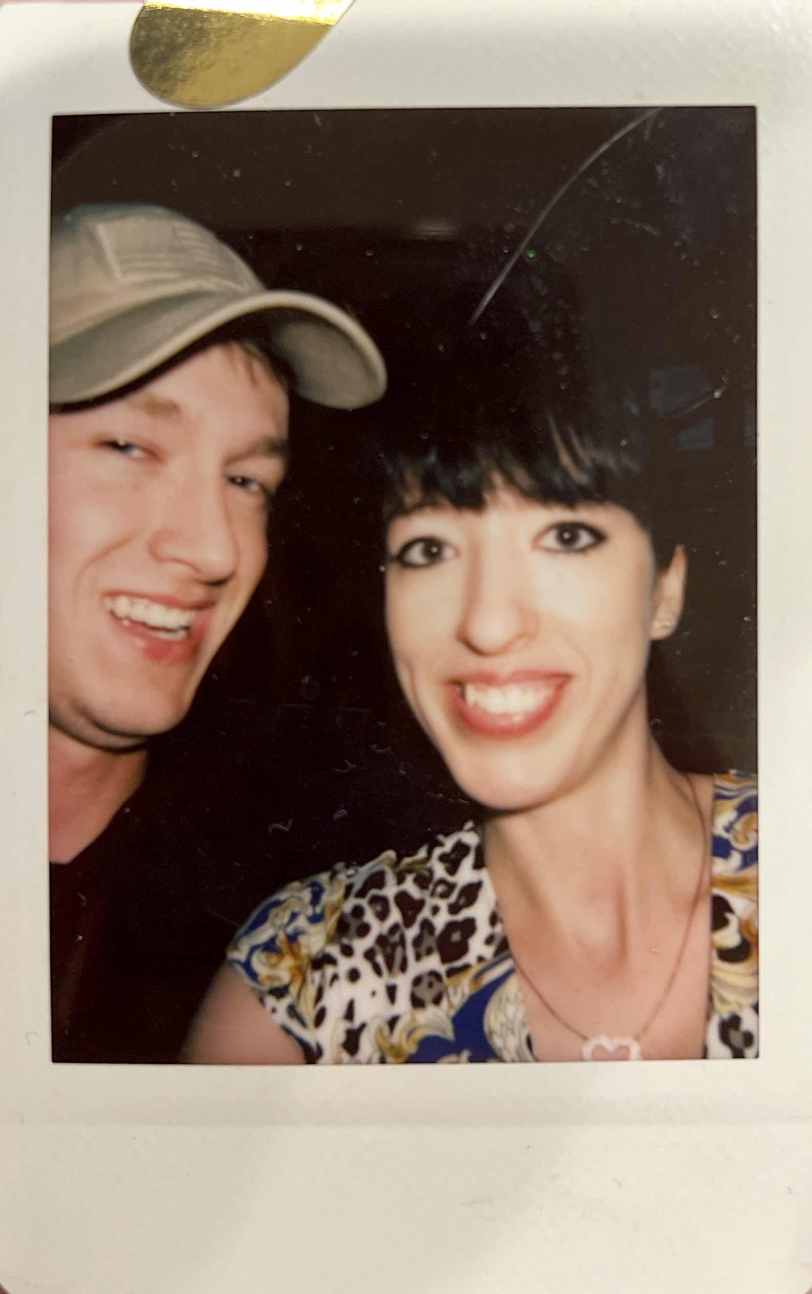 A man wearing a cap and a woman in a patterned dress smiling in a close-up selfie.