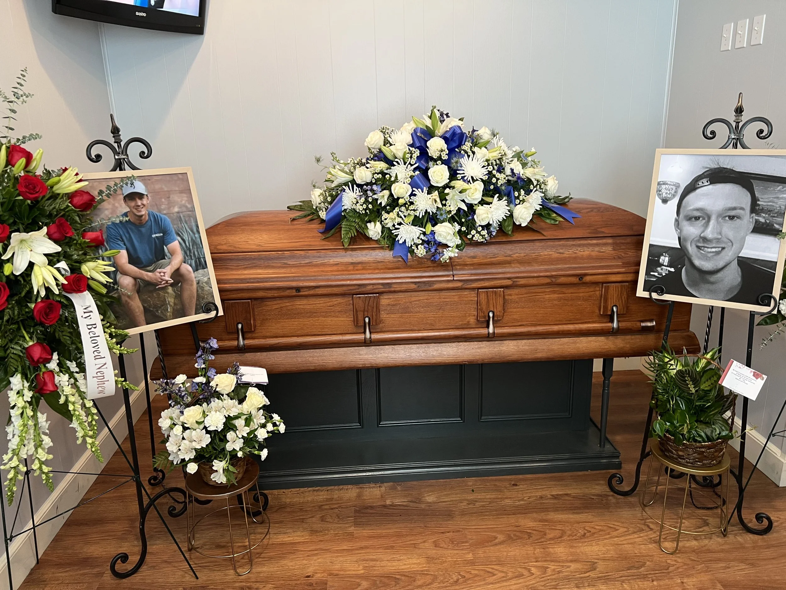 Funeral setting with a wooden coffin with floral arrangements, including white and blue flowers. Two photographs of a young man are displayed on easels. Floral tributes on either side, one with red and white flowers and a ribbon that reads "My Beloved Nephew."