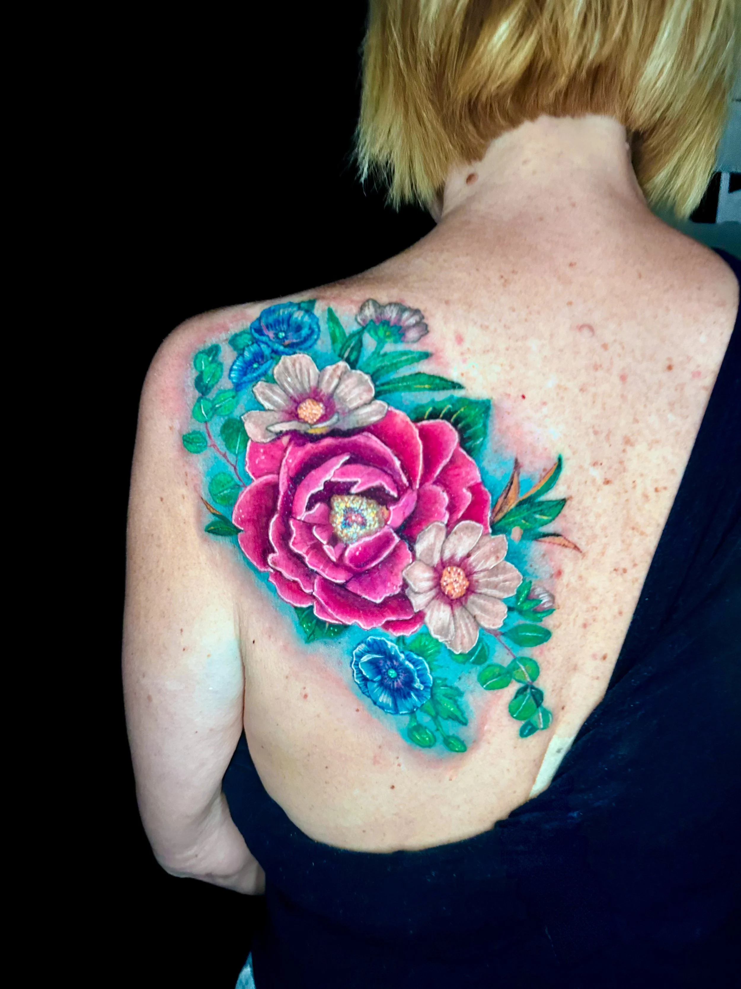 Colorful floral tattoo on person's shoulder with large pink flower, smaller white and blue flowers, and green leaves.
