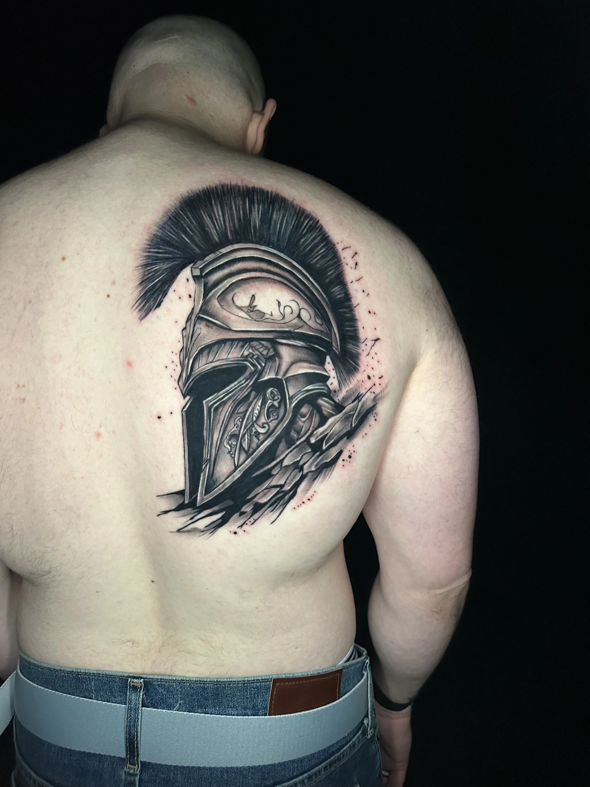 A detailed tattoo of a Greek or Roman helmet with a plume, located on the upper back and shoulder of a person with light skin, wearing jeans.
