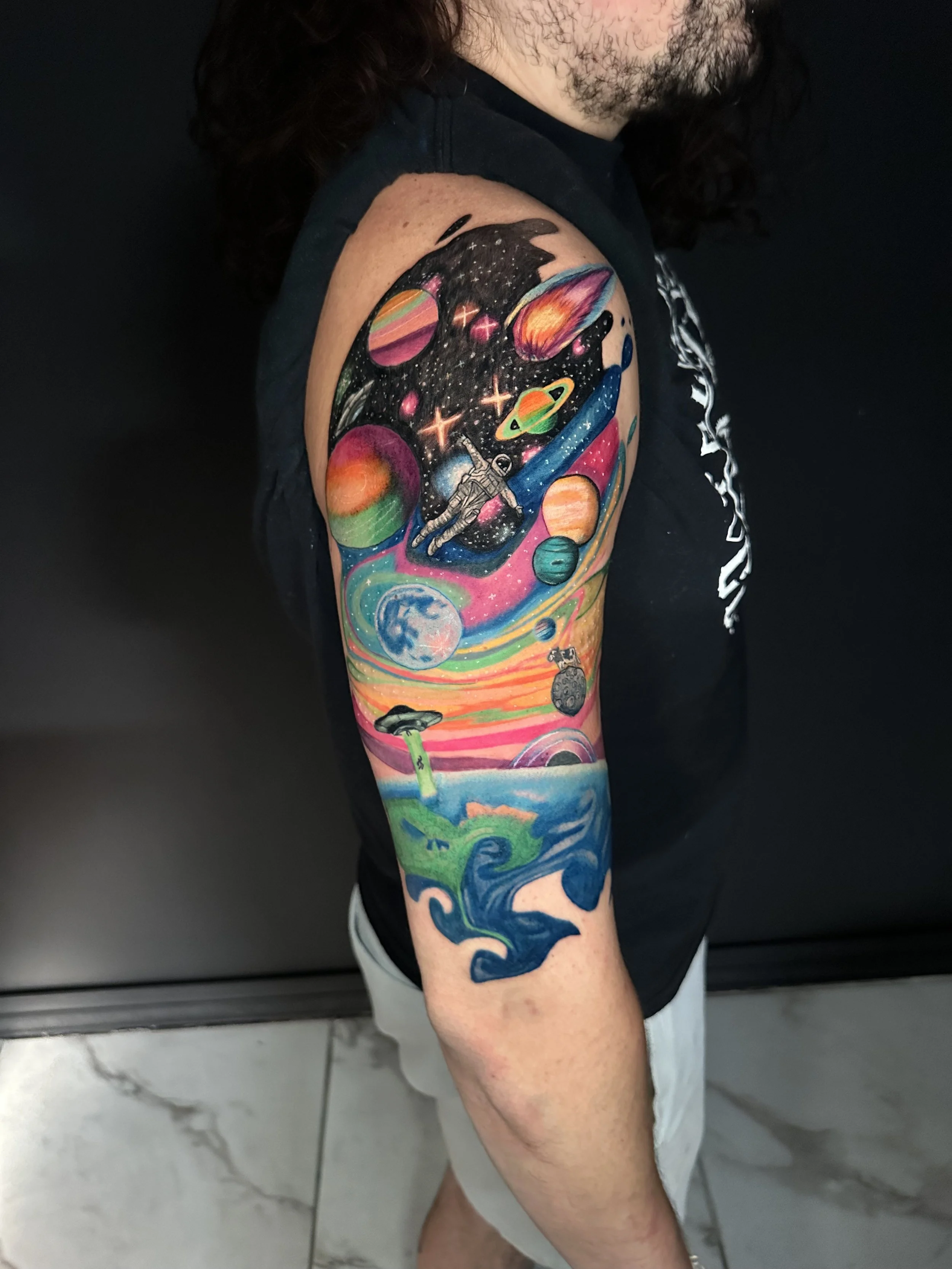 Colorful tattoo sleeve featuring planets, an astronaut, a UFO, a whale, and a vibrant cosmic scene with planets, stars, and a rainbow-style sky.