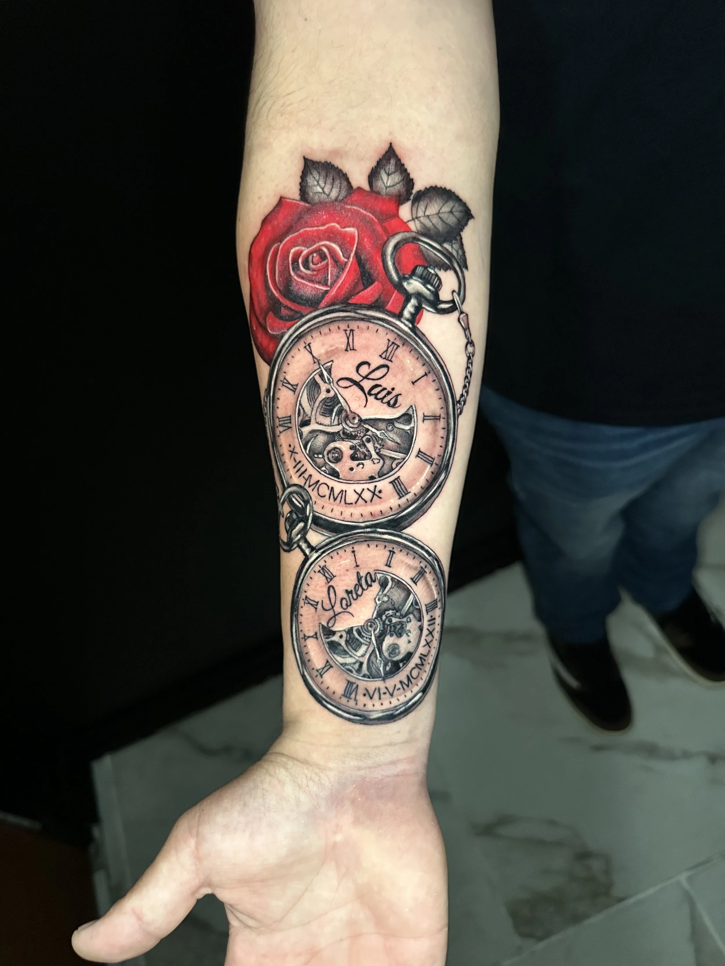 Tattoo of a red rose with green leaves, with two pocket watches beneath it, one with the name 'Lais' and the other 'Laura', and the other with the names 'Lais' and 'Laura'.
