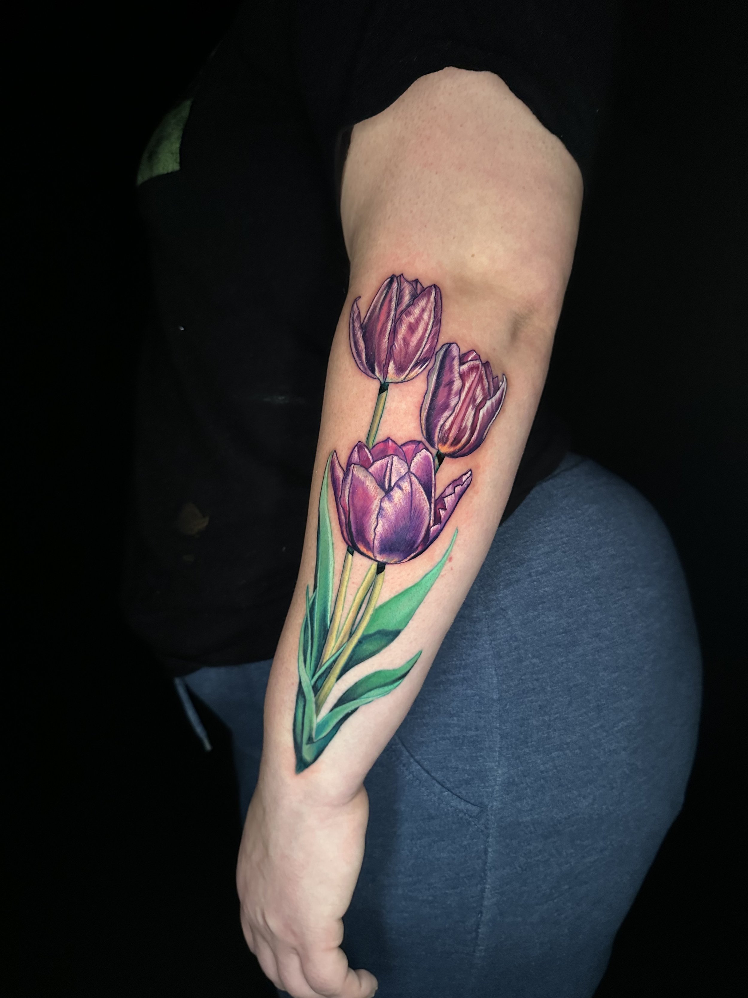 Colorful watercolor-style tattoo of purple tulips and green leaves on a person's forearm.