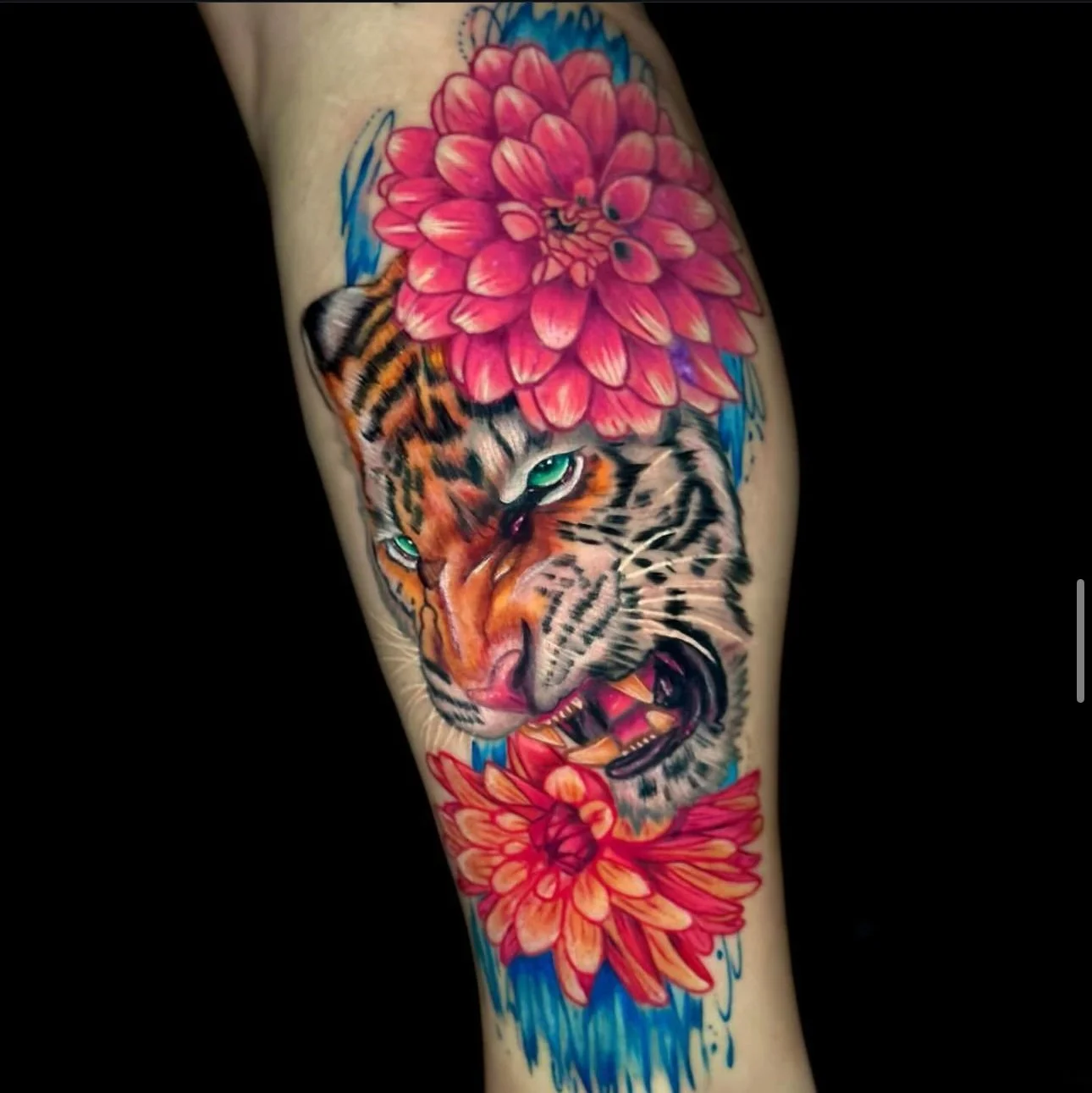 Colorful tattoo of a fierce tiger's face with green eyes, surrounded by large pink flowers and blue brushstroke accents.