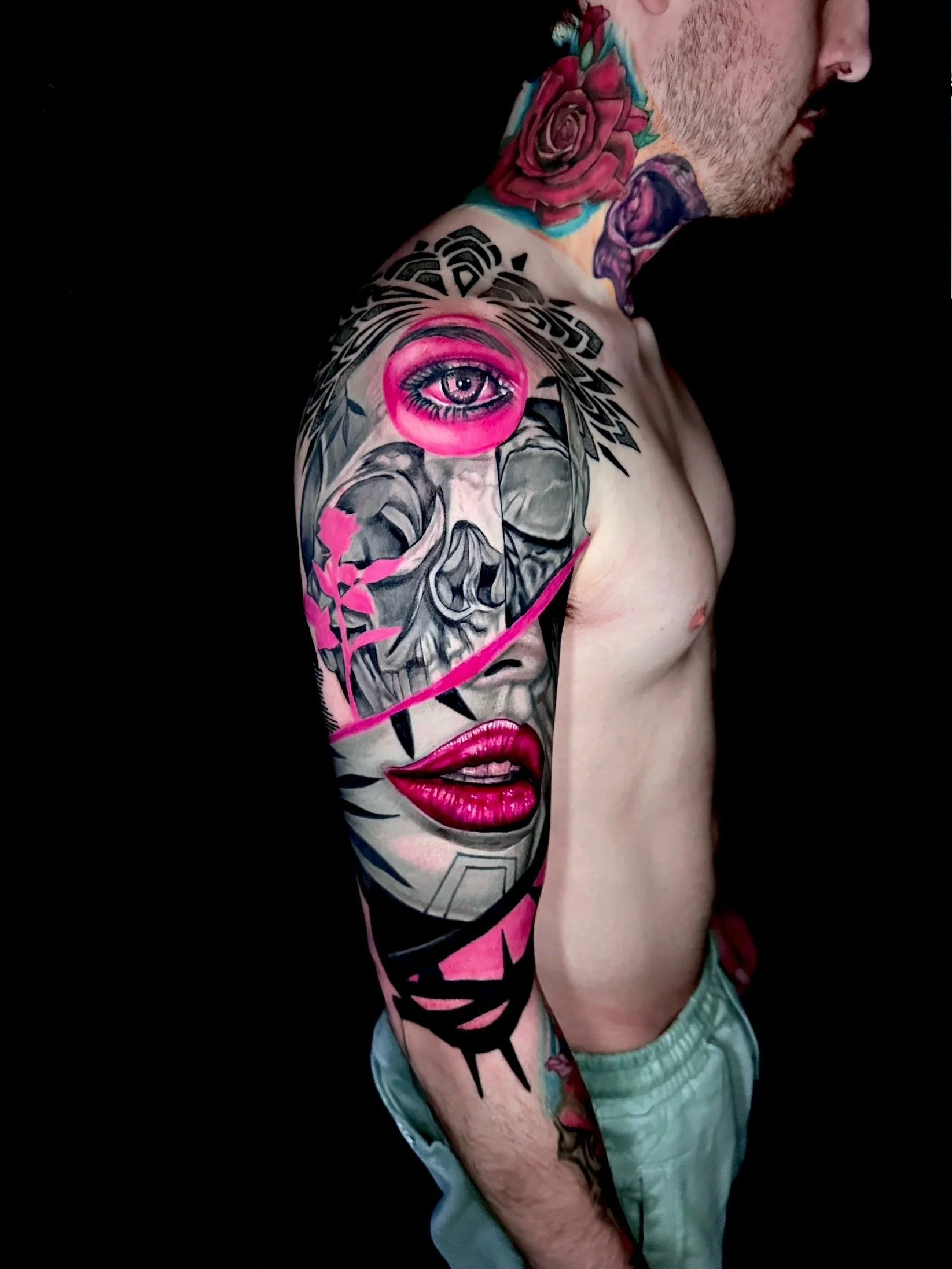 A shirtless man with extensive colorful tattoos on his shoulder, upper arm, and neck, featuring an eye, skull, lips, roses, and abstract designs.