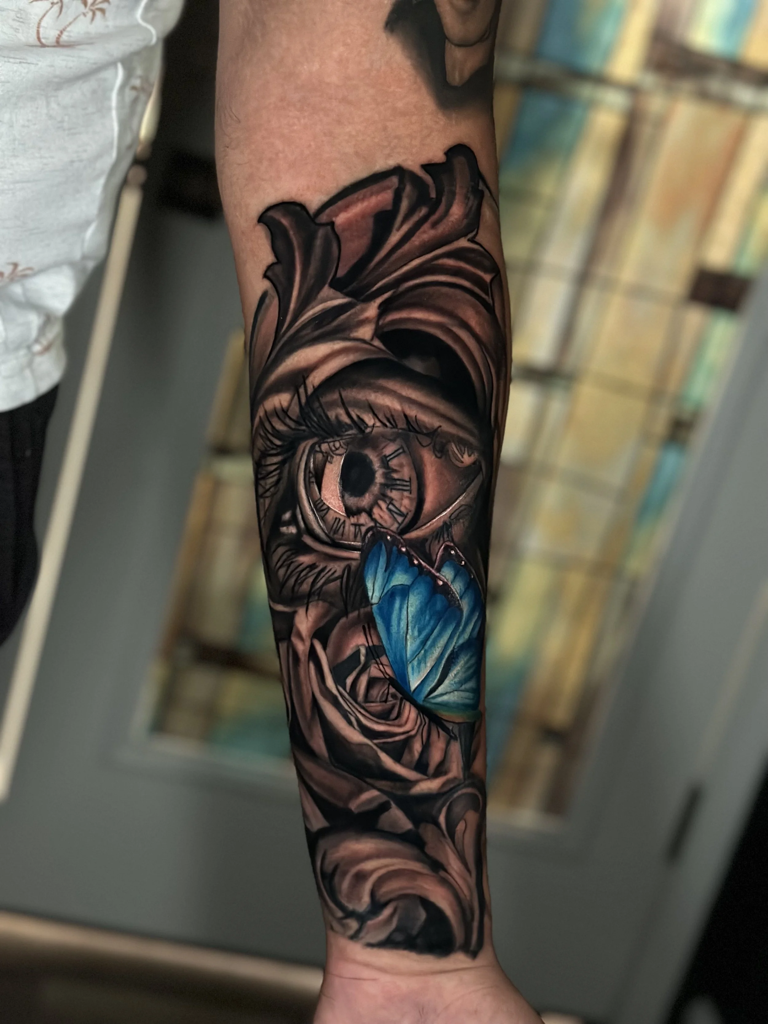 Tattoo of a realistic eye with a clock face in the iris, surrounded by a rose and a blue butterfly on a person's forearm.