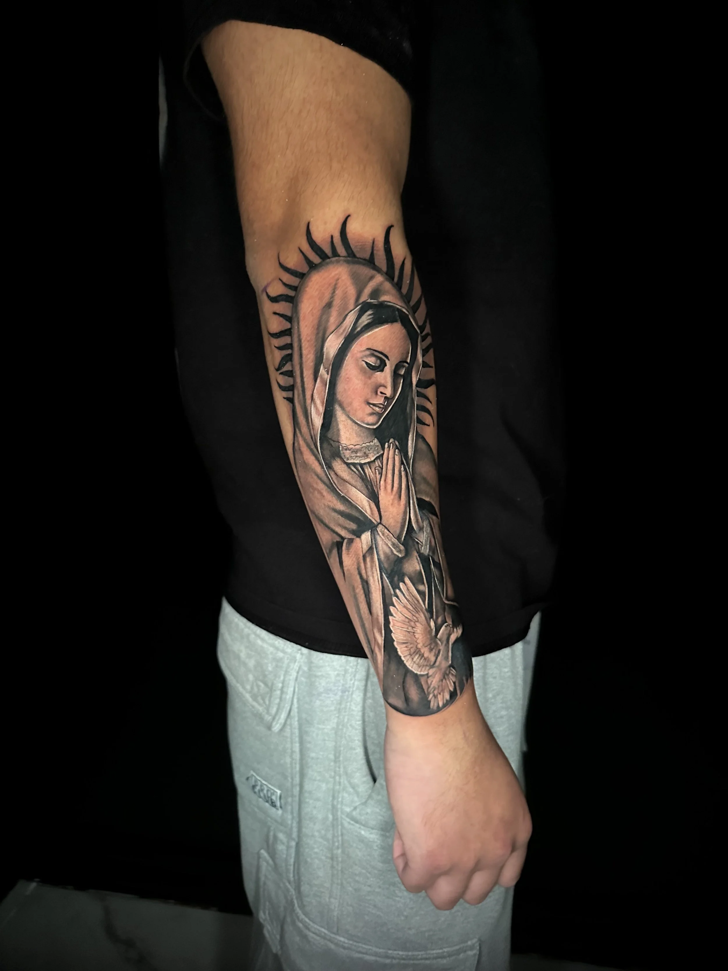 Tattoo of the Virgin Mary on a person's arm, featuring her praying with a halo and a dove.