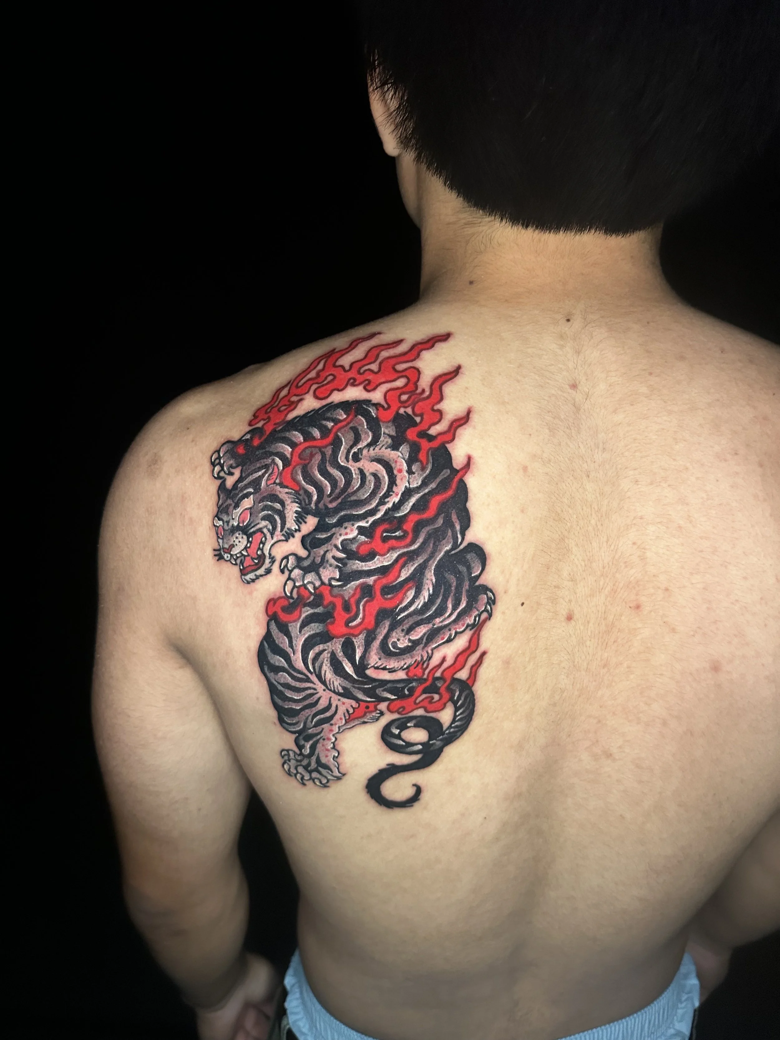 Tattoo of a fierce tiger surrounded by red flames on a person's shoulder and upper back, with short black hair and light skin.