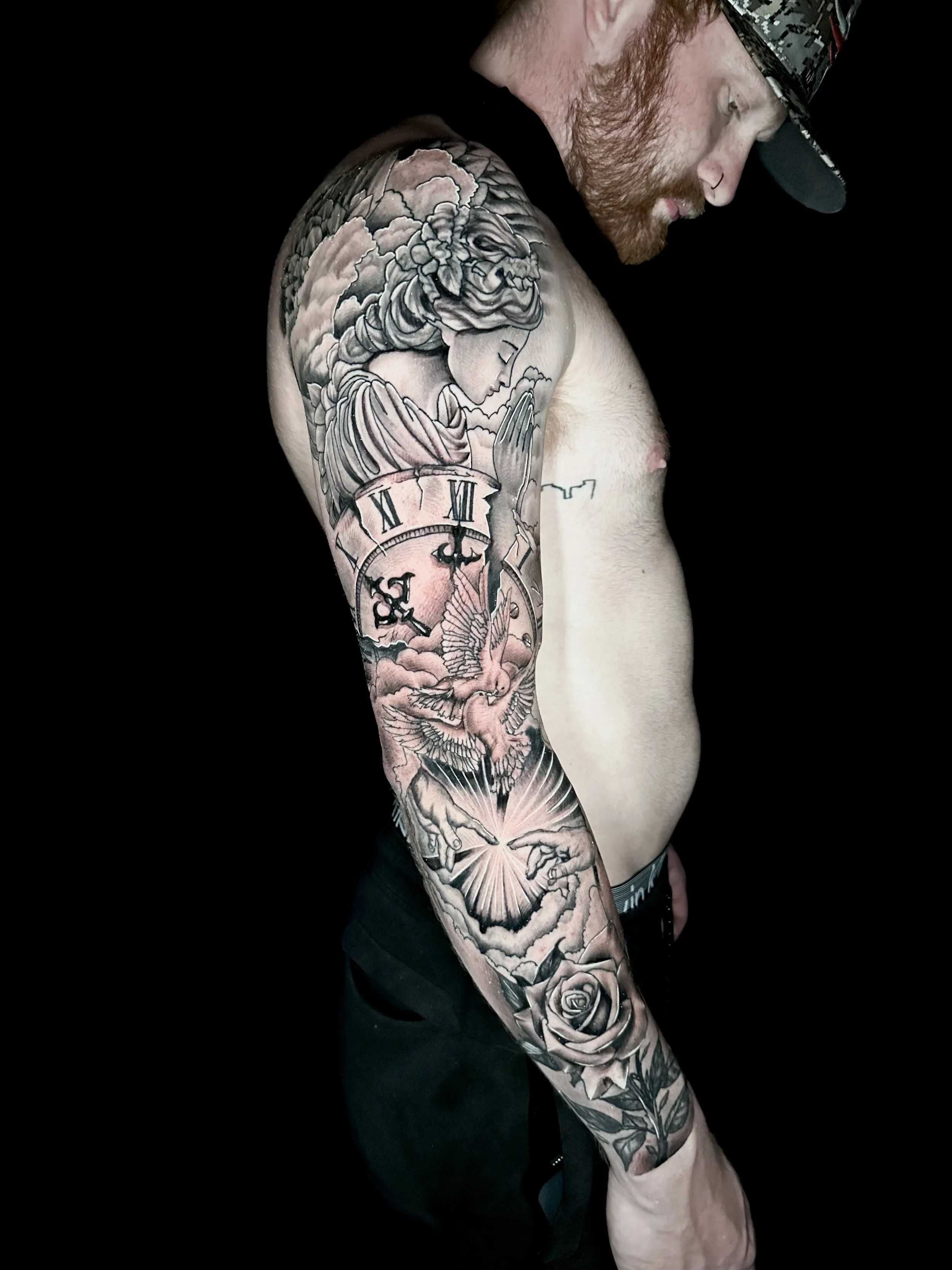 A man with a beard and hat shows a detailed black and gray tattoo sleeve featuring a woman, clock, bird, hands, and roses on his left arm.