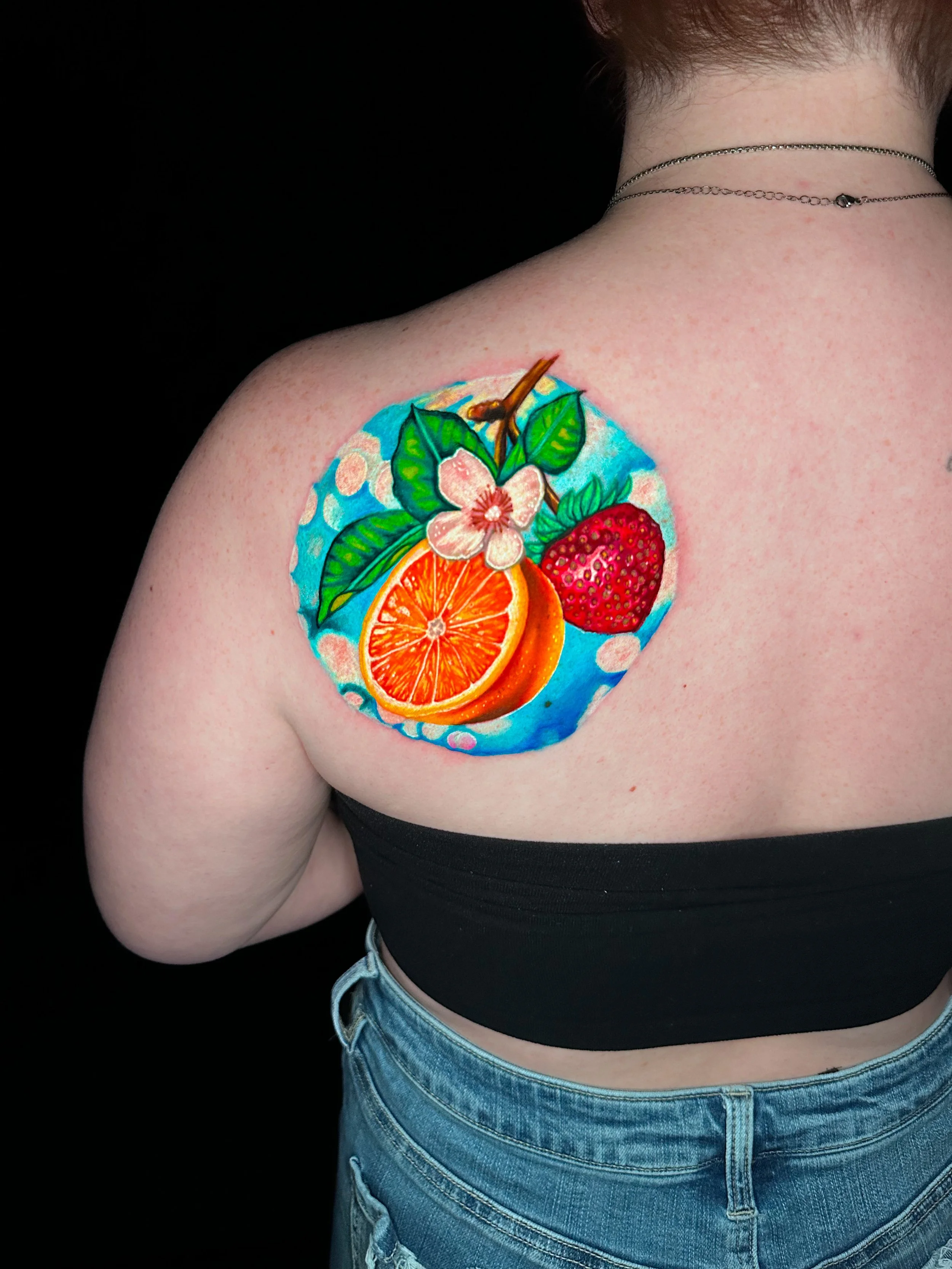 Colorful tattoo of an orange slice, strawberry, and cherry blossom on person's shoulder with blue background