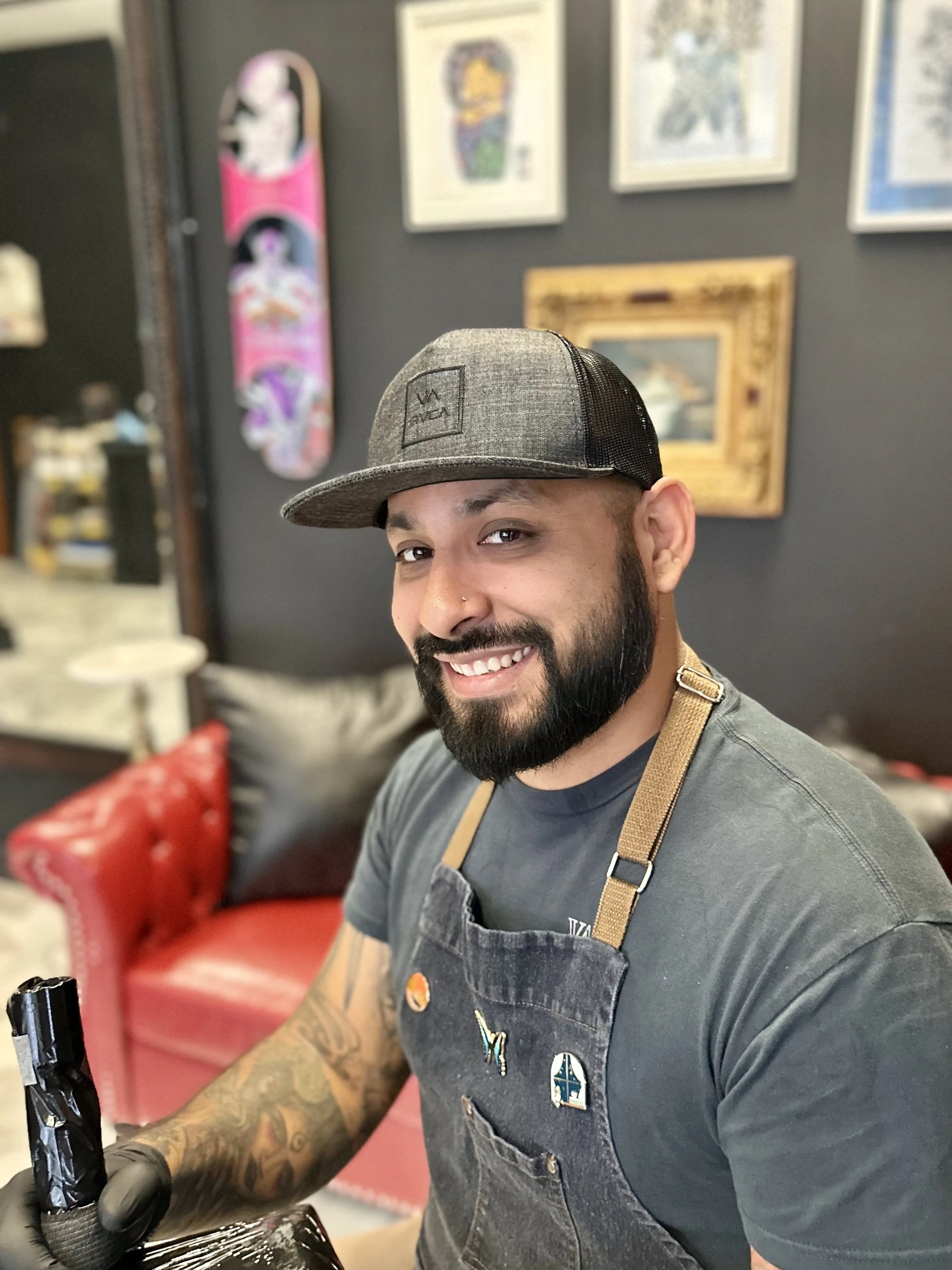 A man with a beard and tattoos on his arm, wearing a gray baseball cap and a dark apron, smiling while working in a tattoo or art studio.