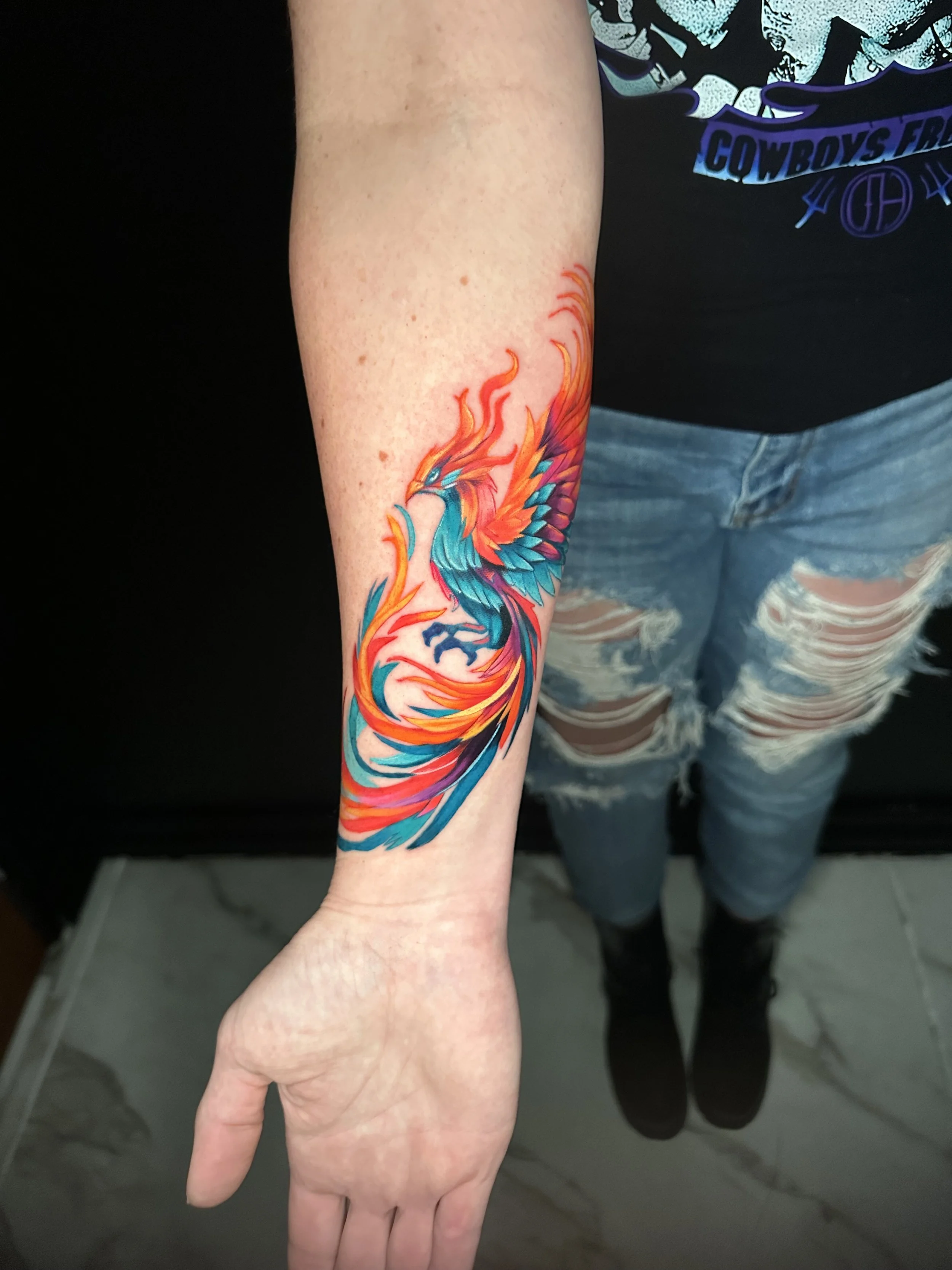 Colorful phoenix tattoo on person's forearm with red, orange, teal, and purple flames.