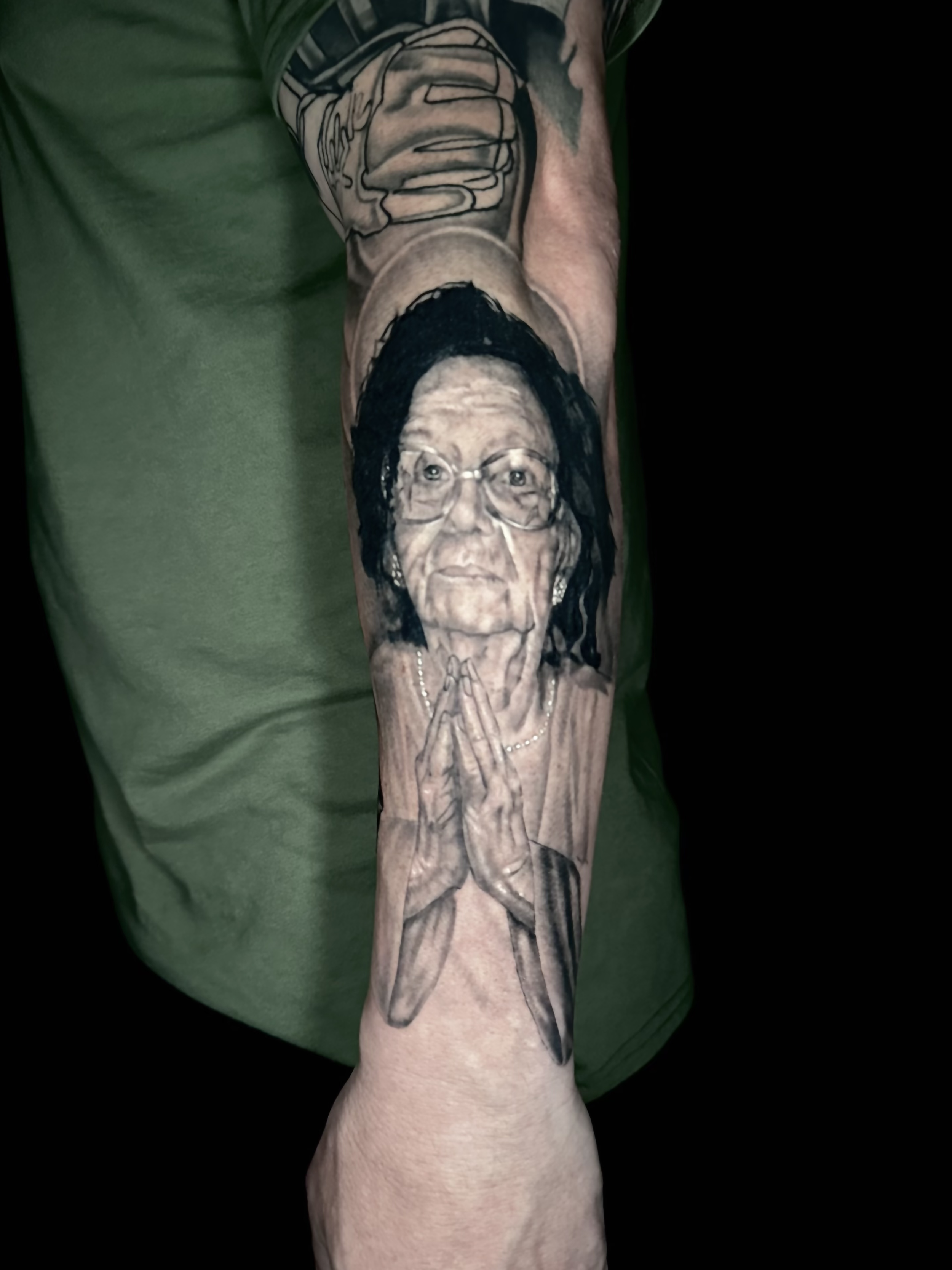 Tattoo of a woman with glasses praying with hands clasped, and sweatshirts above her head on the upper arm.
