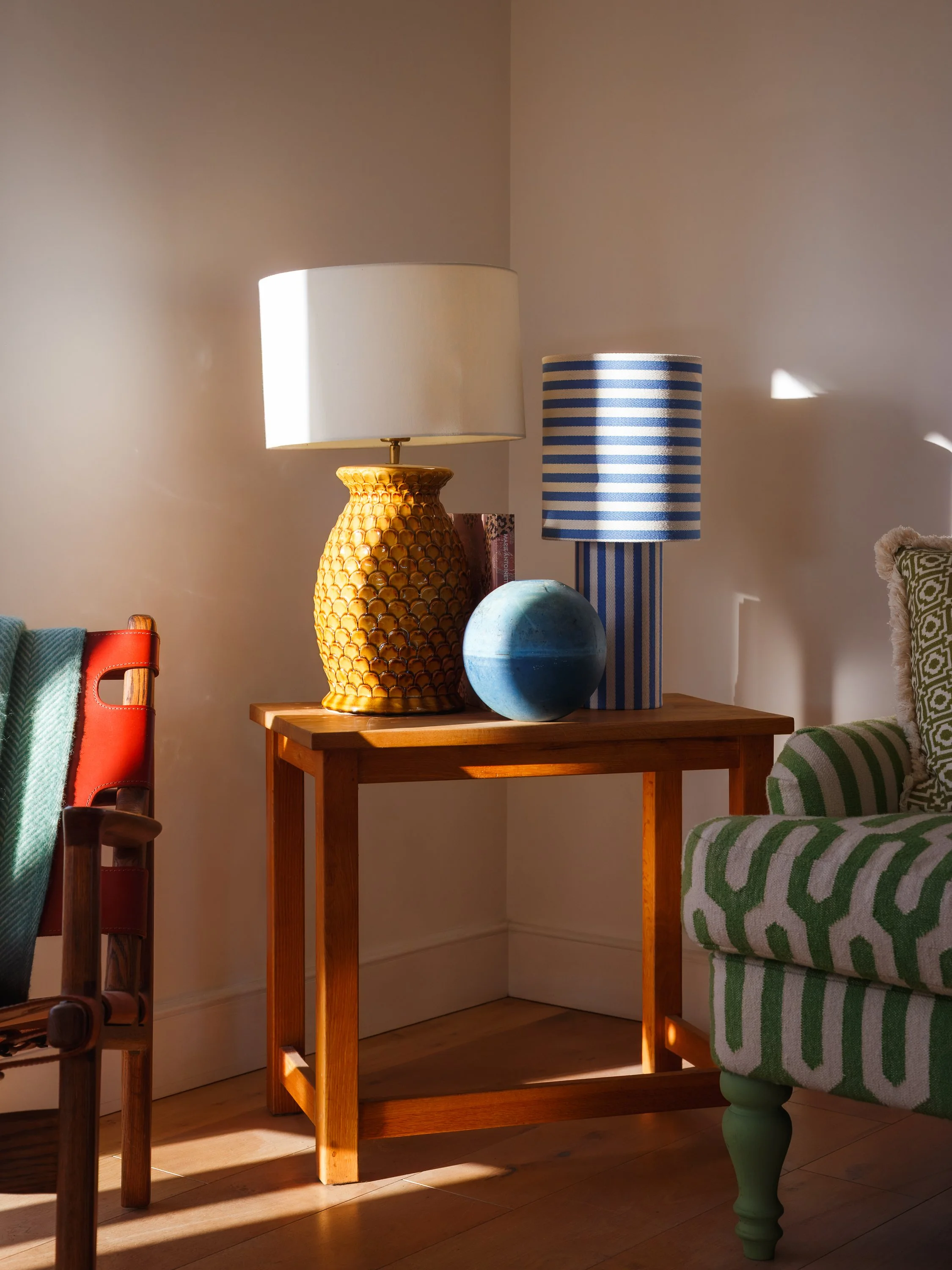 A wooden side table with a yellow textured ceramic lamp, a blue sphere, and a striped blue and white lamp. Part of a green and white patterned chair is visible to the right, and part of a red and brown chair is on the left. The setting is a cozy corn