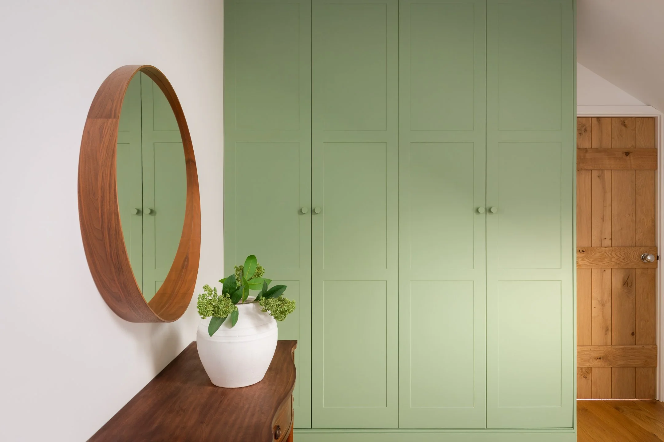 Interior of a room with a wooden dresser holding a potted plant, a large round wooden mirror on the wall, and a wall of green cabinets with a wooden door on the right.