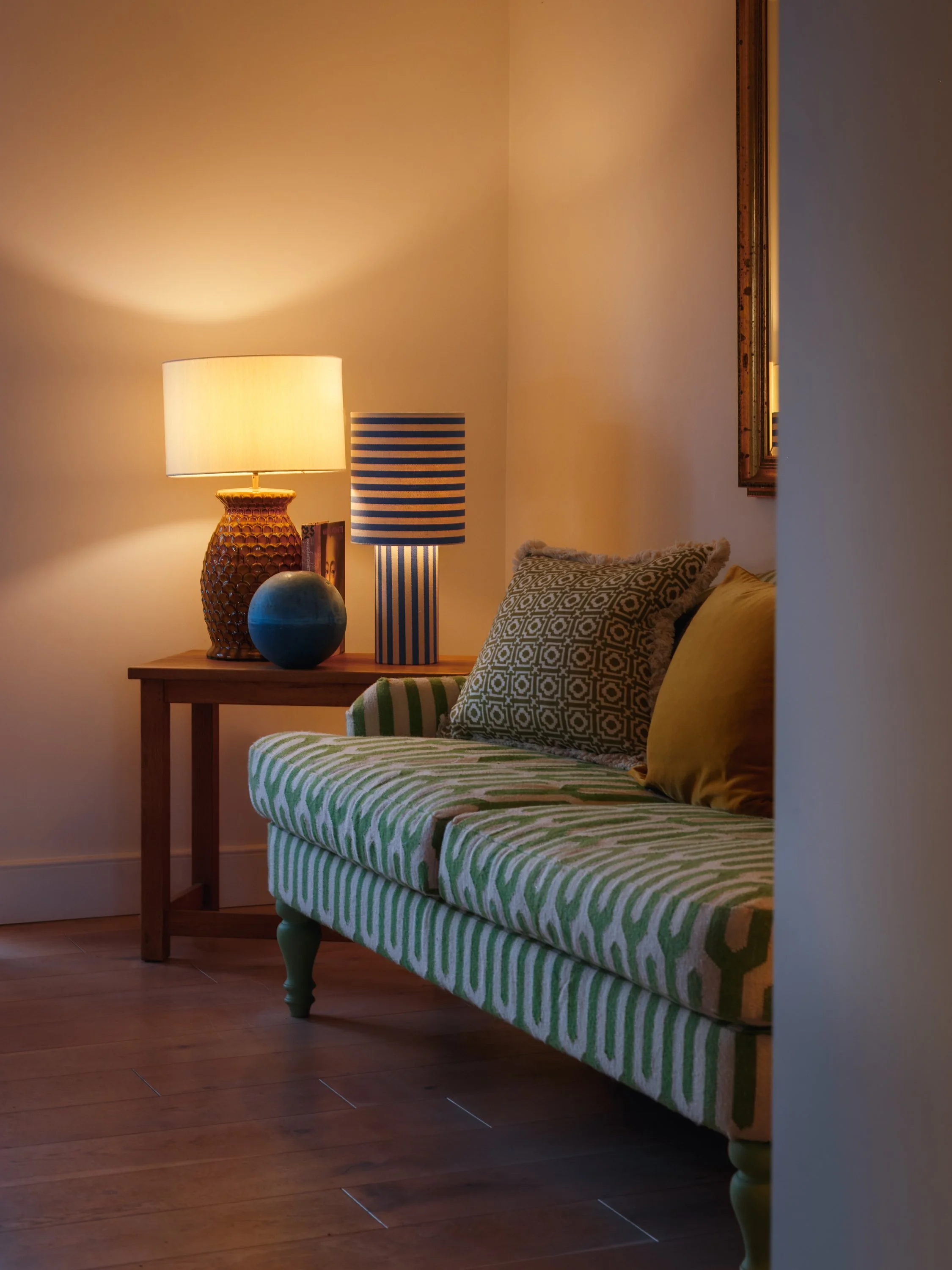 Living room corner with a wooden side table, two table lamps, a decorative vase, and striped and geometric patterned pillows on a green and beige patterned sofa.