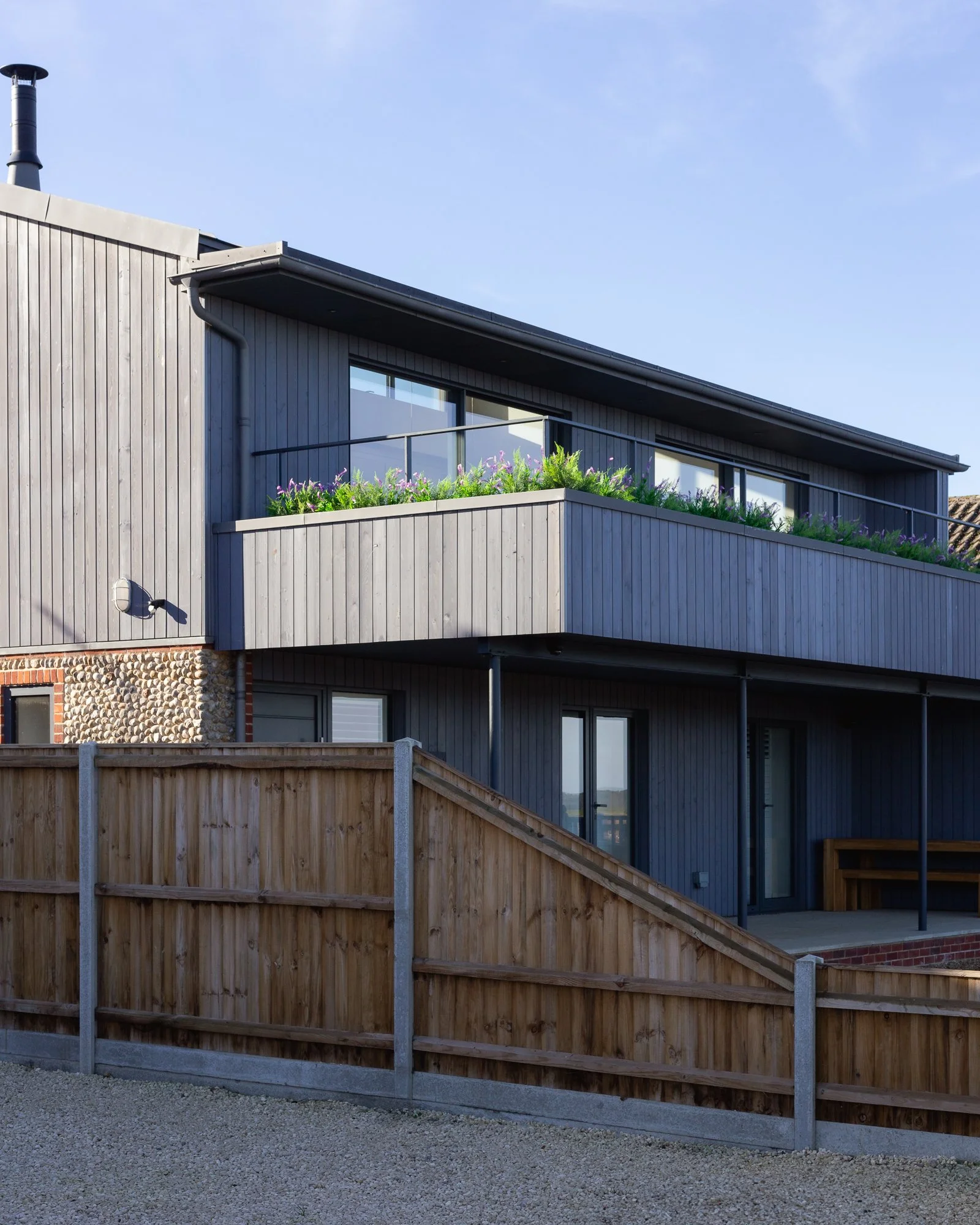Modern multi-story house with gray wooden exteriors and glass sliding doors, featuring a balcony with purple flowers, surrounded by a wooden fence under a clear blue sky.