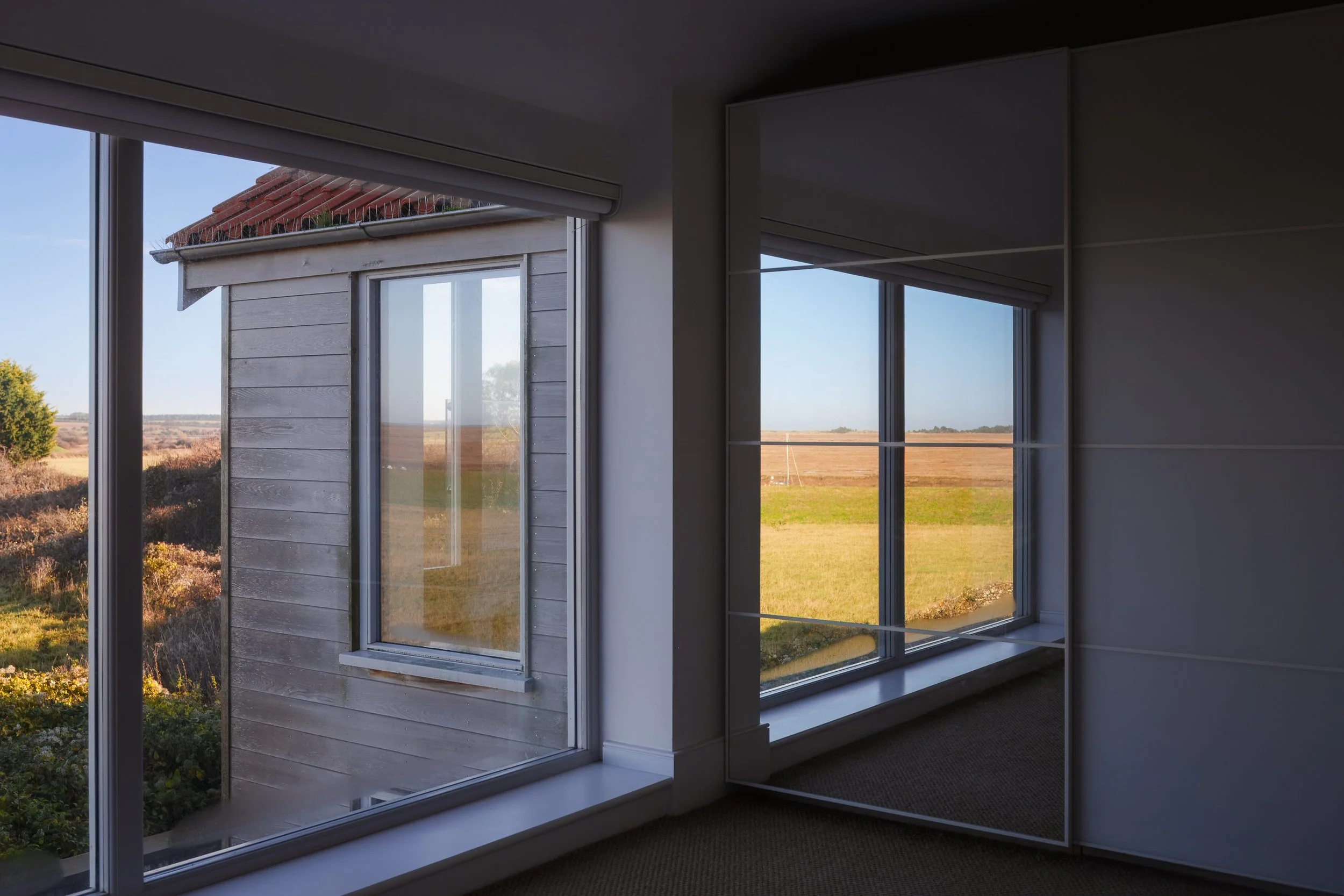 Interior view of a room with large windows looking out onto a grassy field and a wooden building with a red roof