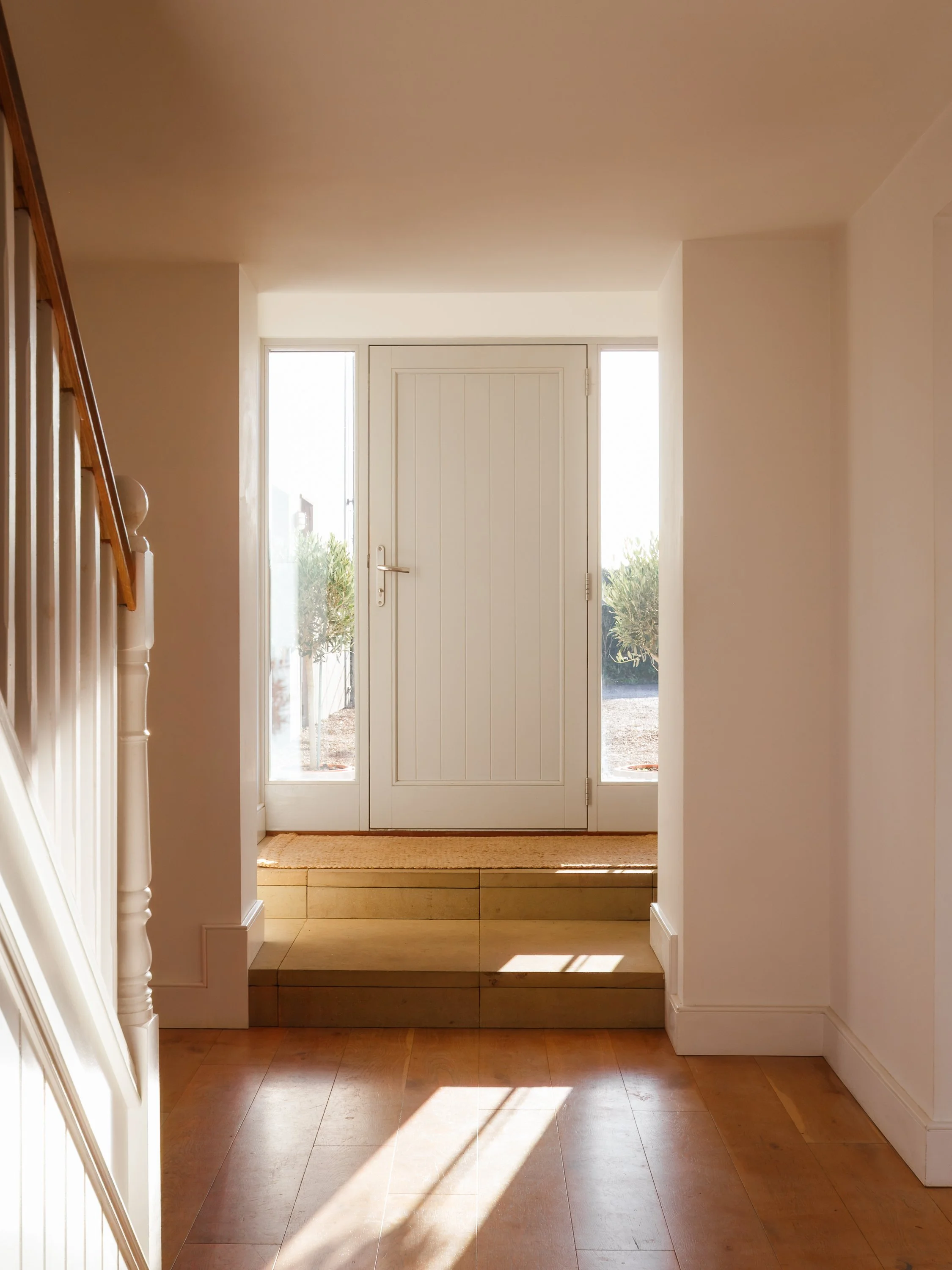 Interior view of a bright foyer with a white door and glass panels, wooden stairs on the left, and sunlight streaming onto the wooden floor.