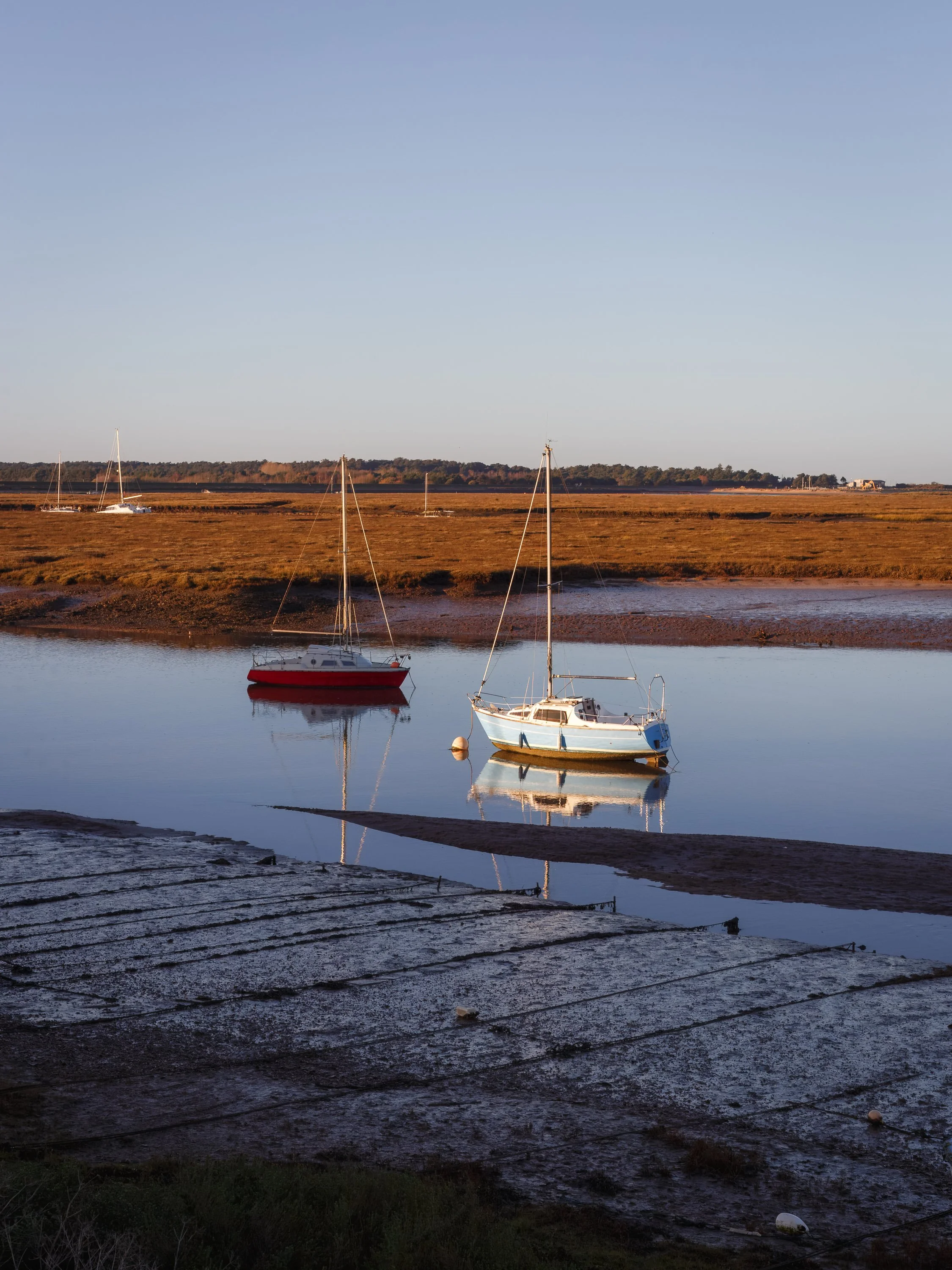 Boats anchored in a calm body of water during low tide, with a clear sky and a marshy landscape in the background.