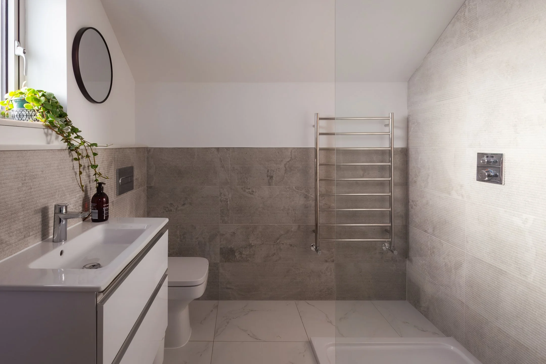 Modern bathroom with small sink vanity, toilet, wall-mounted heated towel rail, window with potted plant, and textured wall tiles.