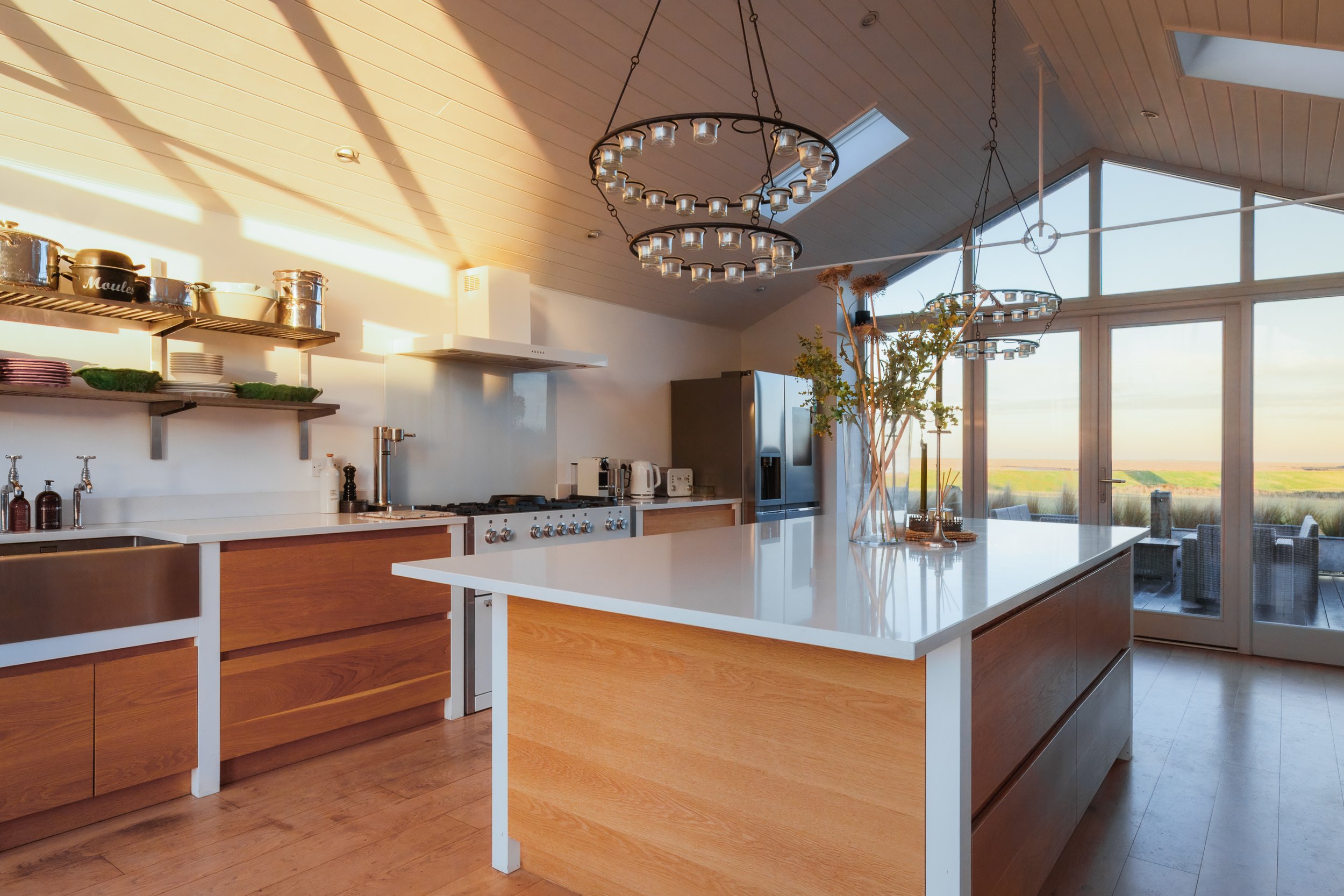 Modern kitchen with wooden cabinets, white countertops, and large windows letting in natural light. There's a kitchen island with white surface, a stainless steel refrigerator, and a dining outside on a patio with outdoor seating. Decorative dishes, 