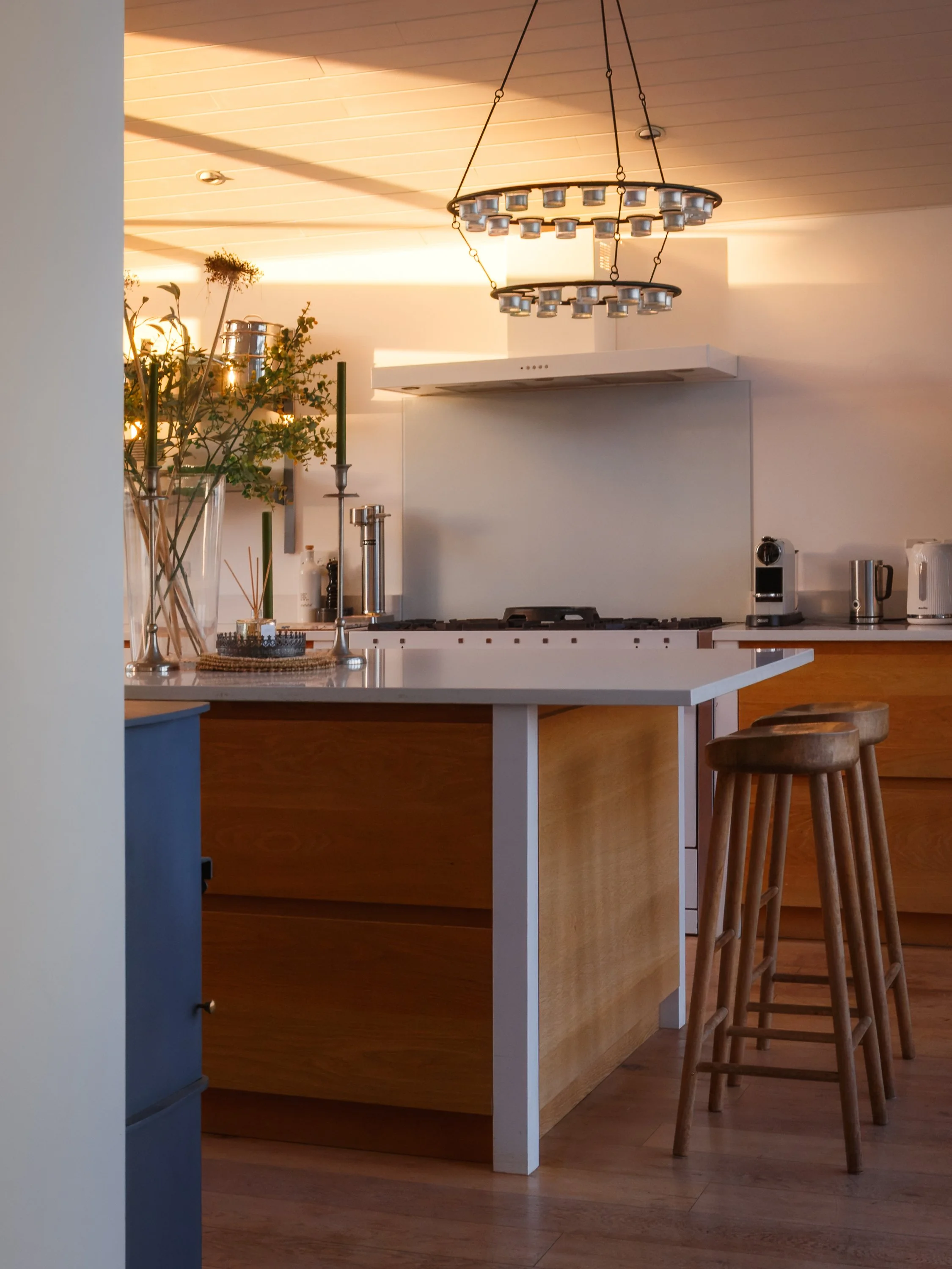 A modern kitchen with a white countertop, wooden cabinets, and stools. There is a chandelier hanging from the ceiling, plants, and appliances on the counter.