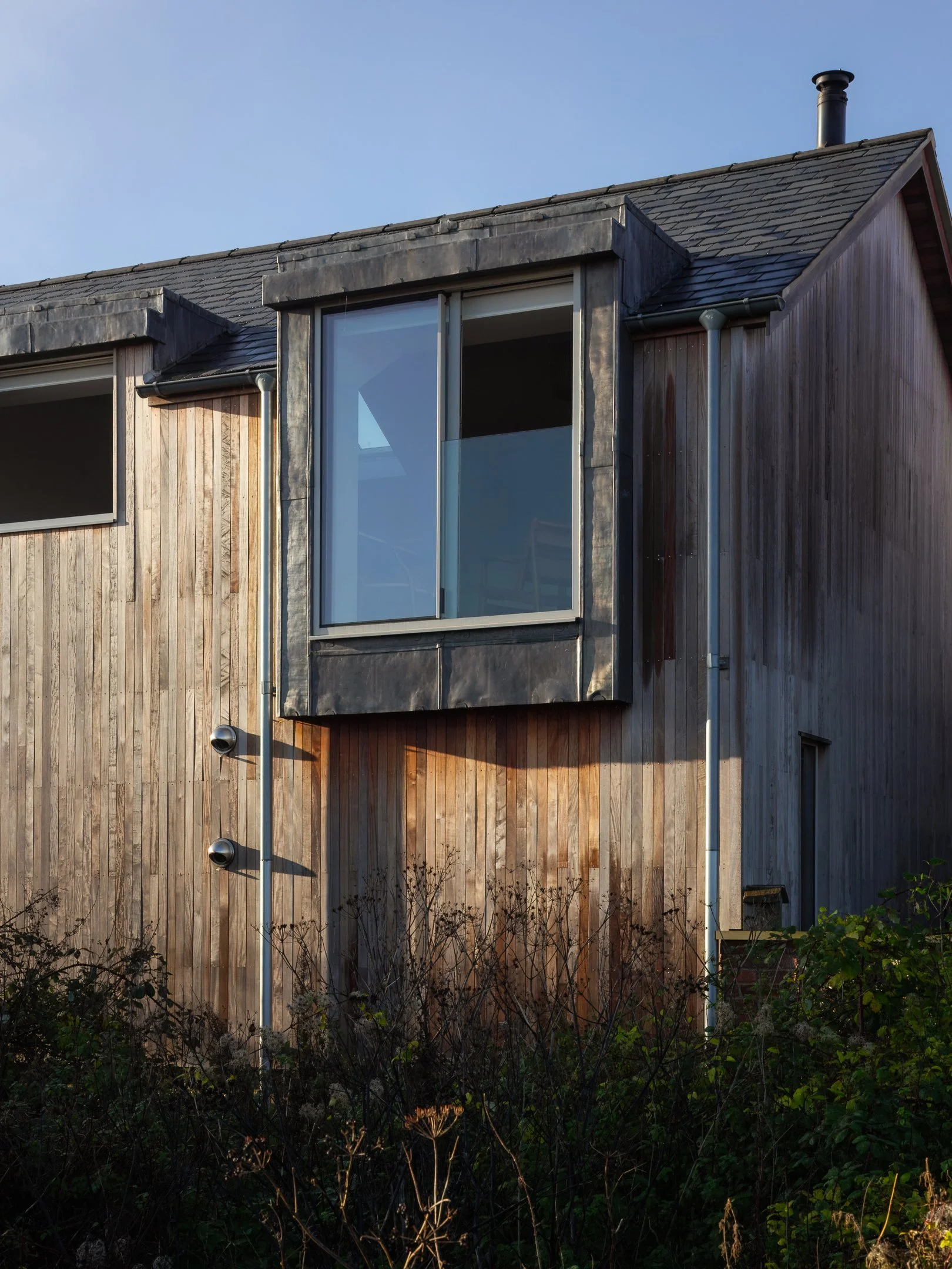 Exterior view of a modern house with wooden siding, large glass window, and a rain gutter system, under a clear blue sky.