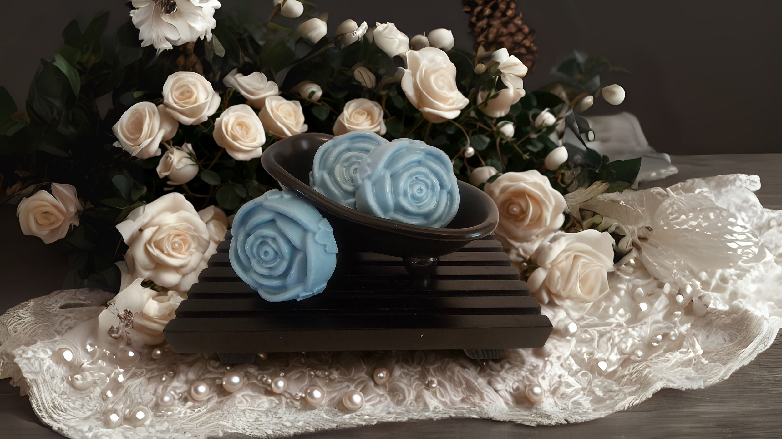 A floral arrangement of white roses, white berries, and green leaves in the background. In the foreground, a black dish with three light blue, swirled soap bars placed on a black slatted surface, surrounded by pearl-like beads and white lace fabric.