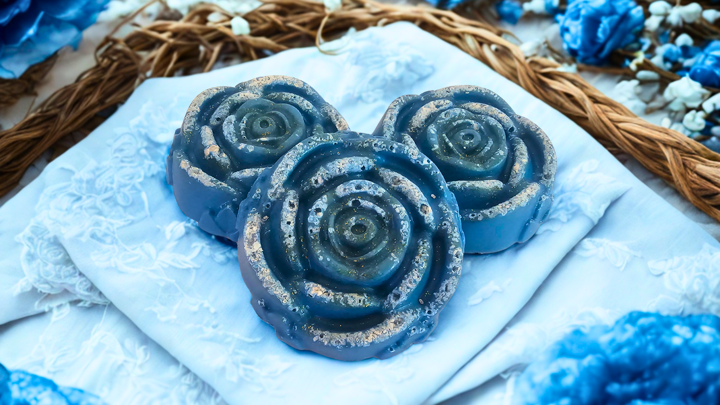Three blue soap bars shaped like roses placed on white fabric with lace, surrounded by other blue and white flowers and a woven basket.