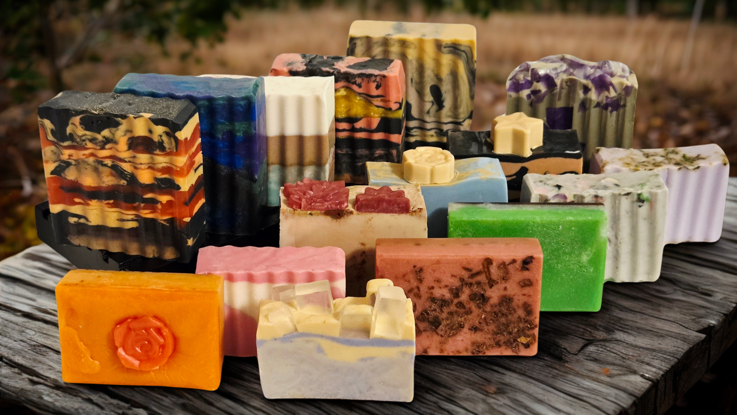 A variety of colorful handmade soap bars arranged outdoors on a wooden surface with a blurred natural background.