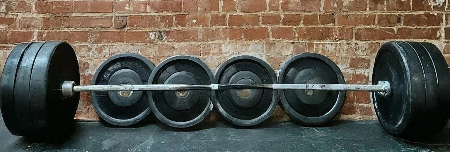 Barbell with weight plates on each end and several smaller weight plates stacked beside it on a black floor against a brick wall.