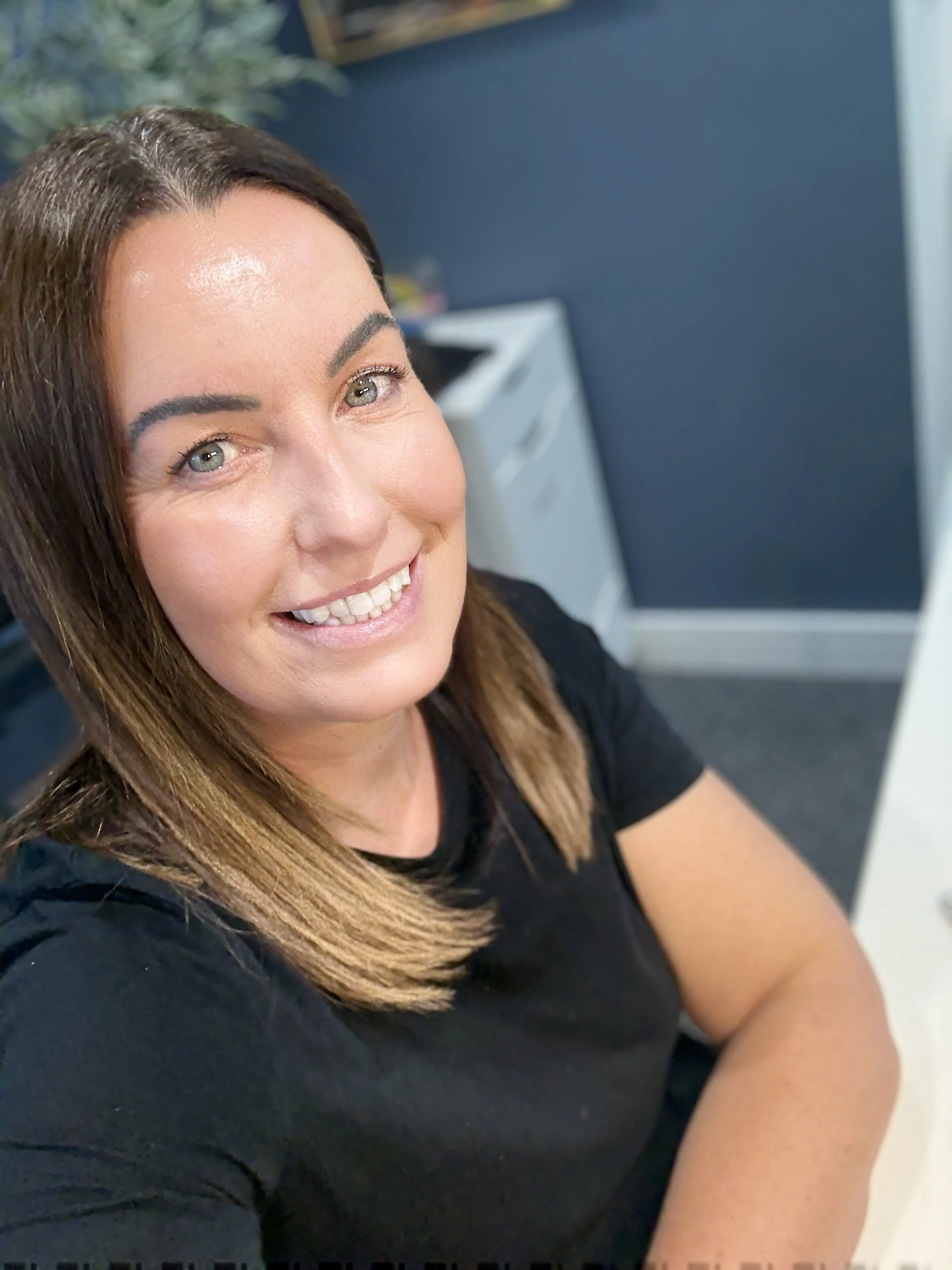 Close-up of a woman with shoulder-length brown hair, smiling, wearing a black shirt, sitting in an office setting.