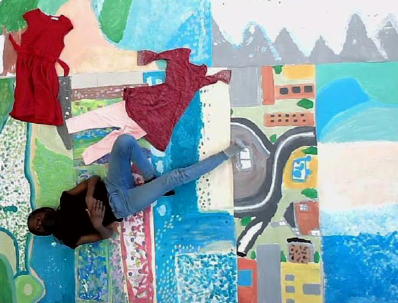 Une femme pose dans une pièce avec un mur peint représentant un paysage urbain avec des montagnes en arrière-plan, des bâtiments, une route, et une rivière, portant un t-shirt noir et un jean, avec deux robes rouges suspendues derrière elle.